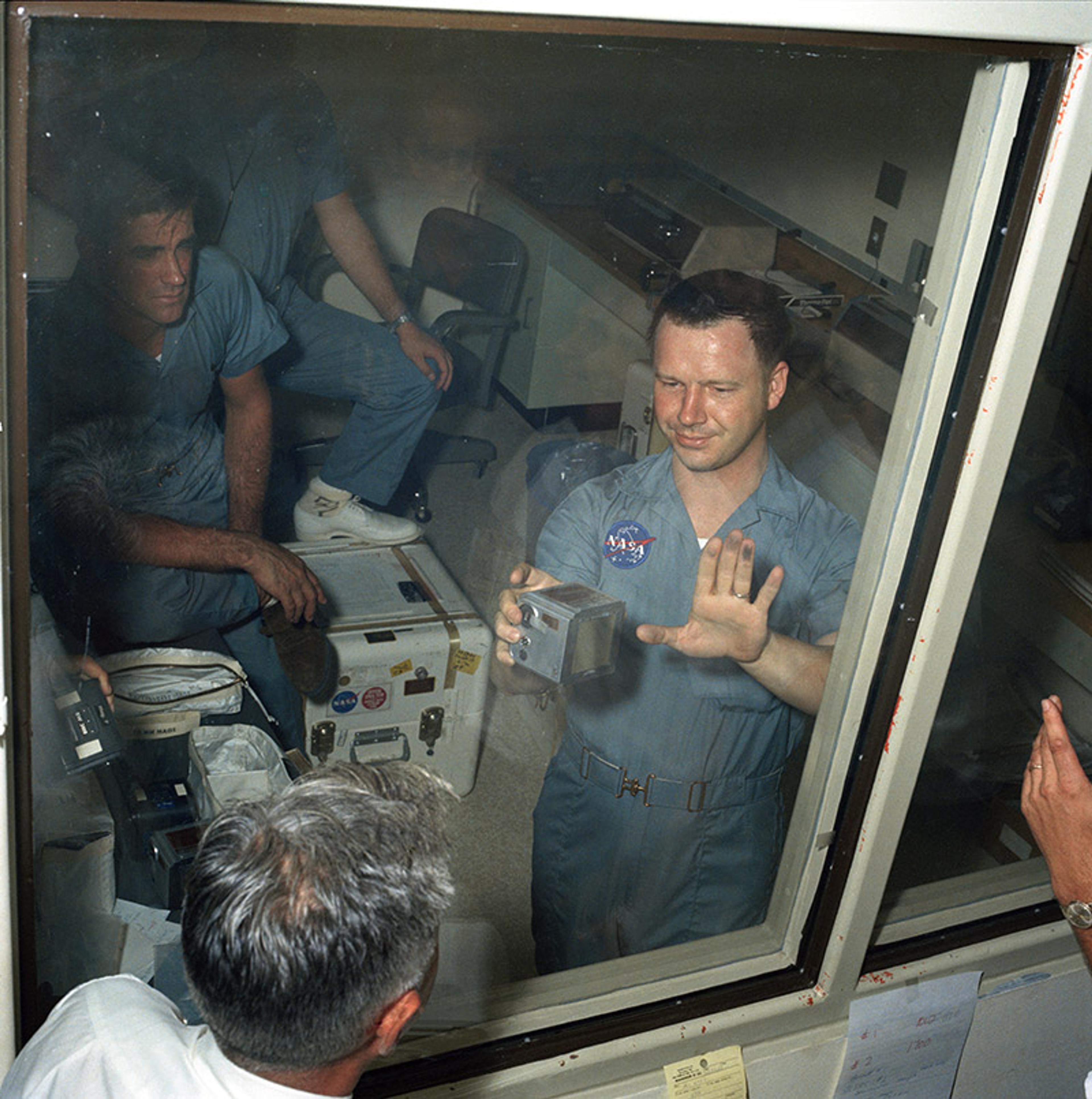 Astronauts in a room, one pressing his hand against a glass window, others sitting behind him in blue uniforms.
