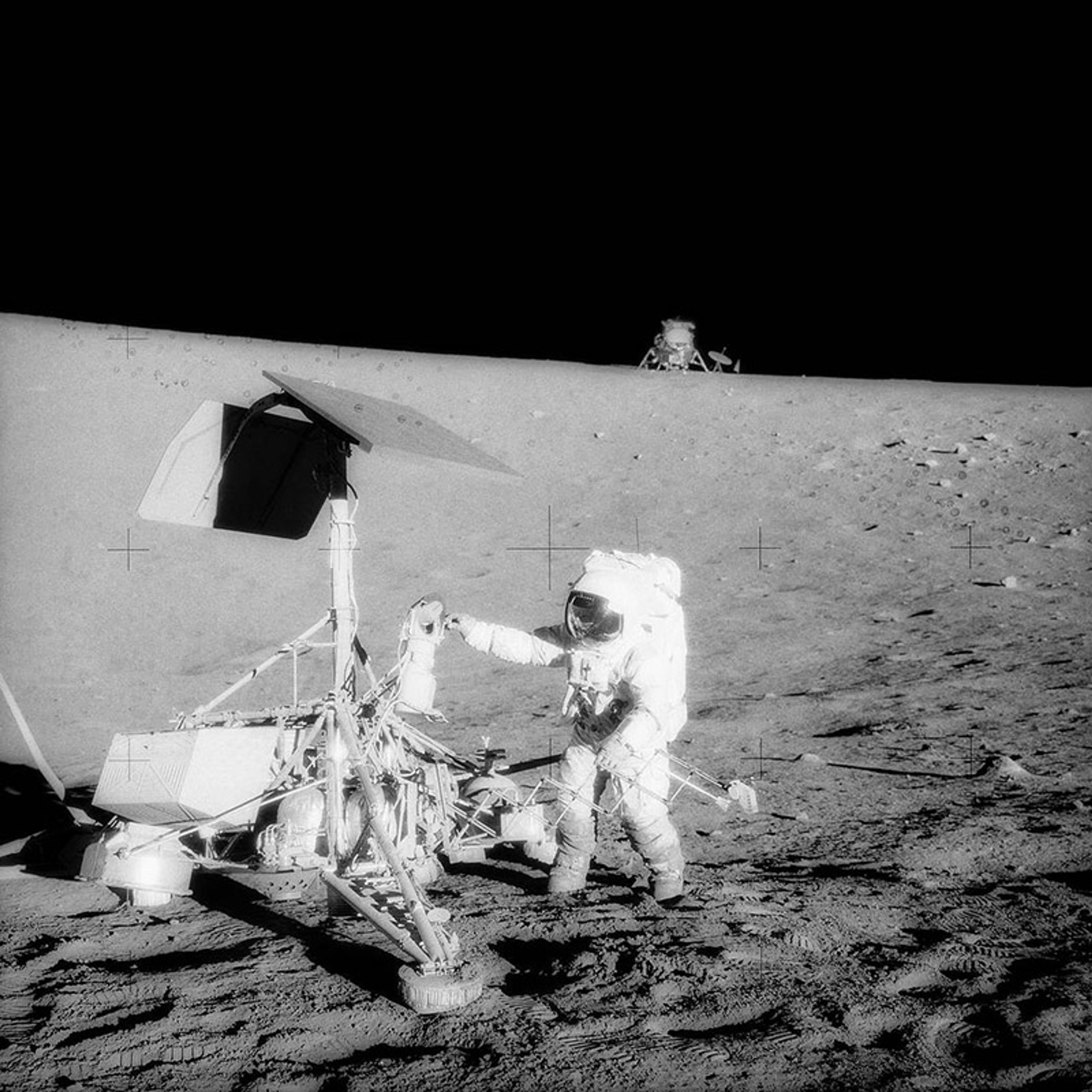 Black-and-white photo of an astronaut examining a lunar lander on the Moon’s surface with a spacecraft in the background.