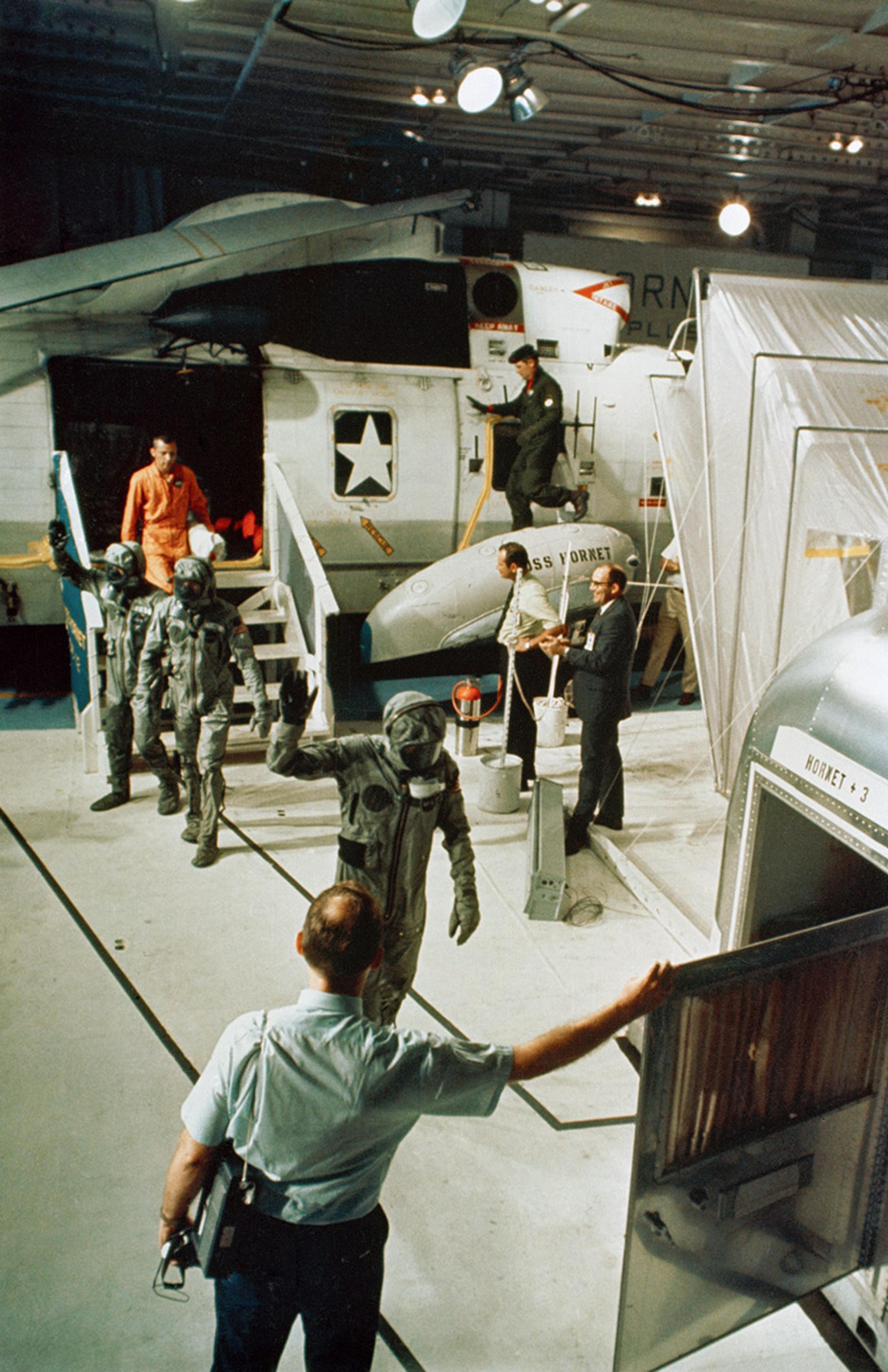 Astronauts in suits exiting a helicopter onto a ship, with several personnel and equipment around them under bright lights.