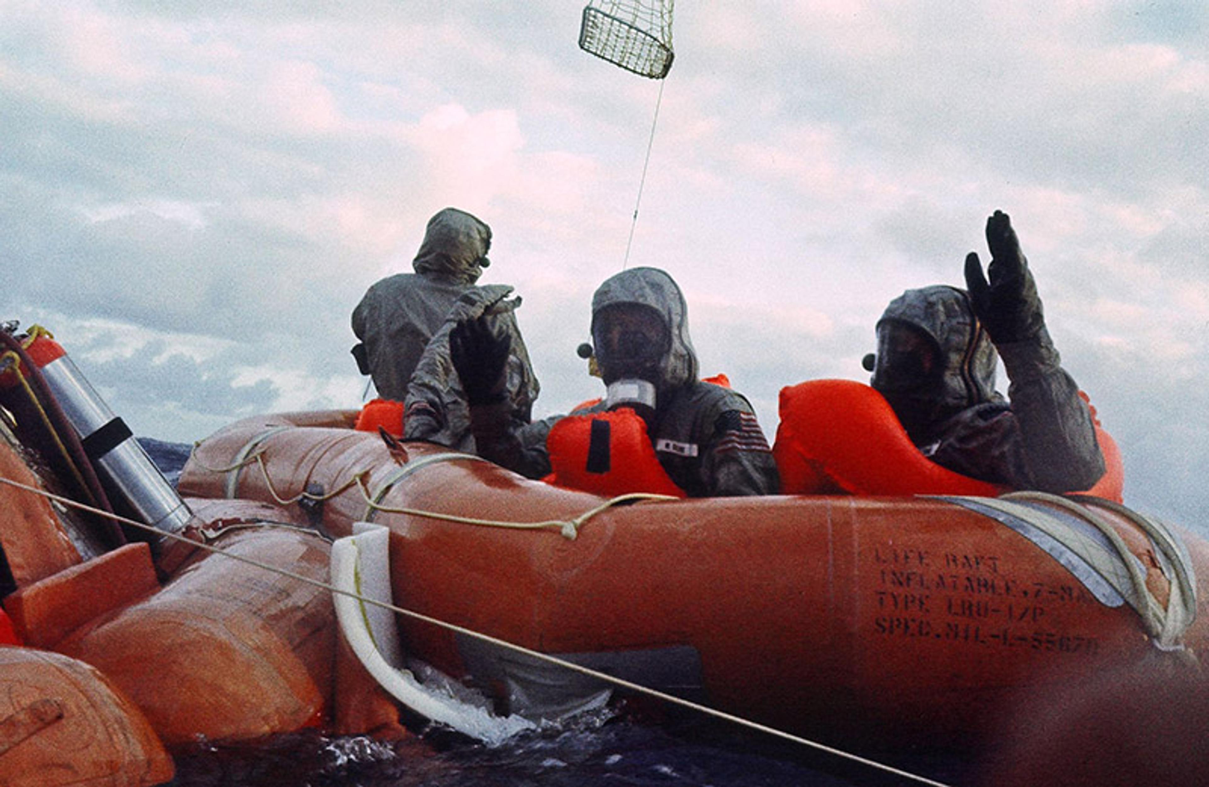 Three astronauts wearing suits in an orange life raft at sea, with a rescue basket overhead and cloudy sky background.