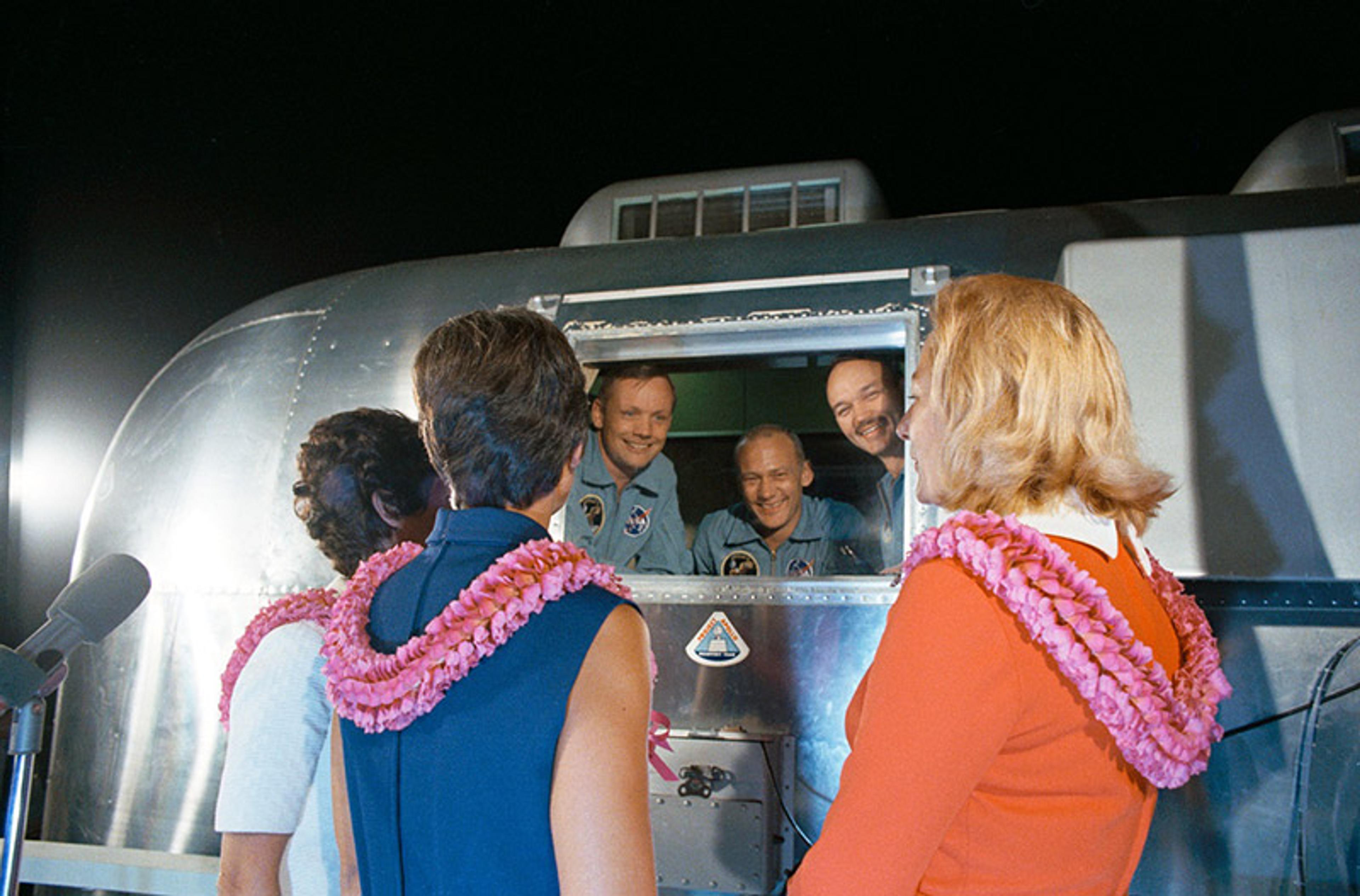 Three astronauts in blue uniforms smiling from a window greeting three women wearing leis outside a space module.