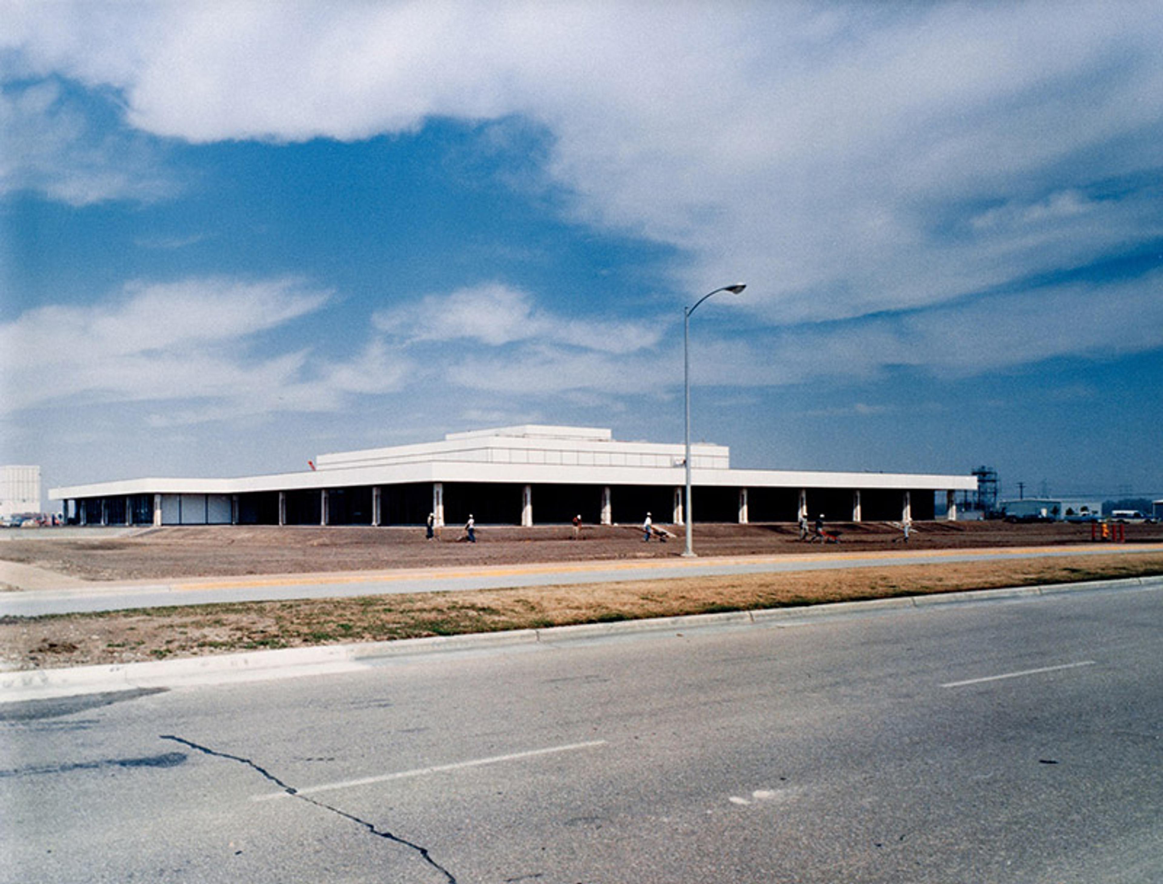 A modern, flat-roofed building under construction, with an open field in the foreground and a cloudy sky above.