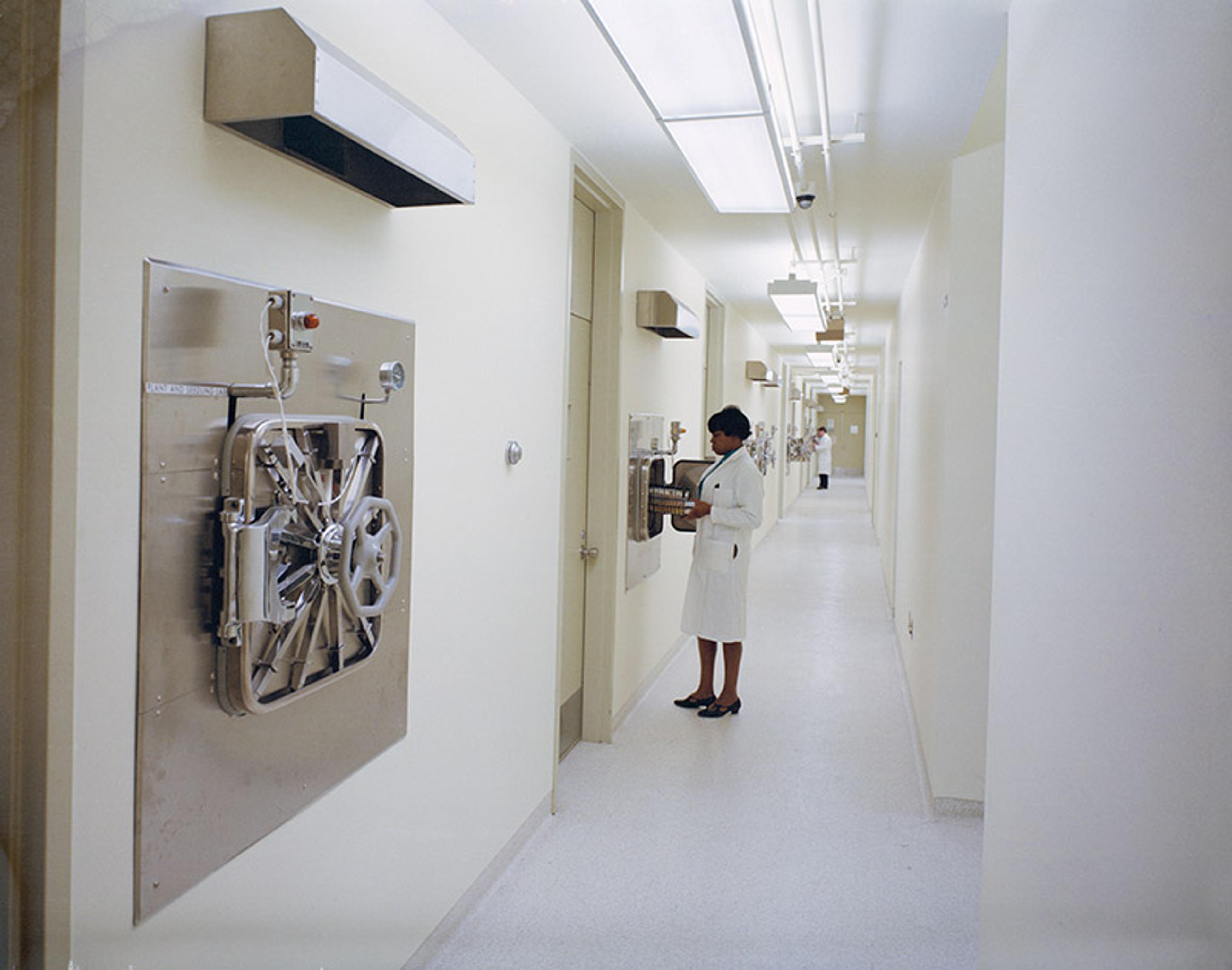 A hospital corridor with medical equipment on walls and a nurse in a white uniform examining a machine.