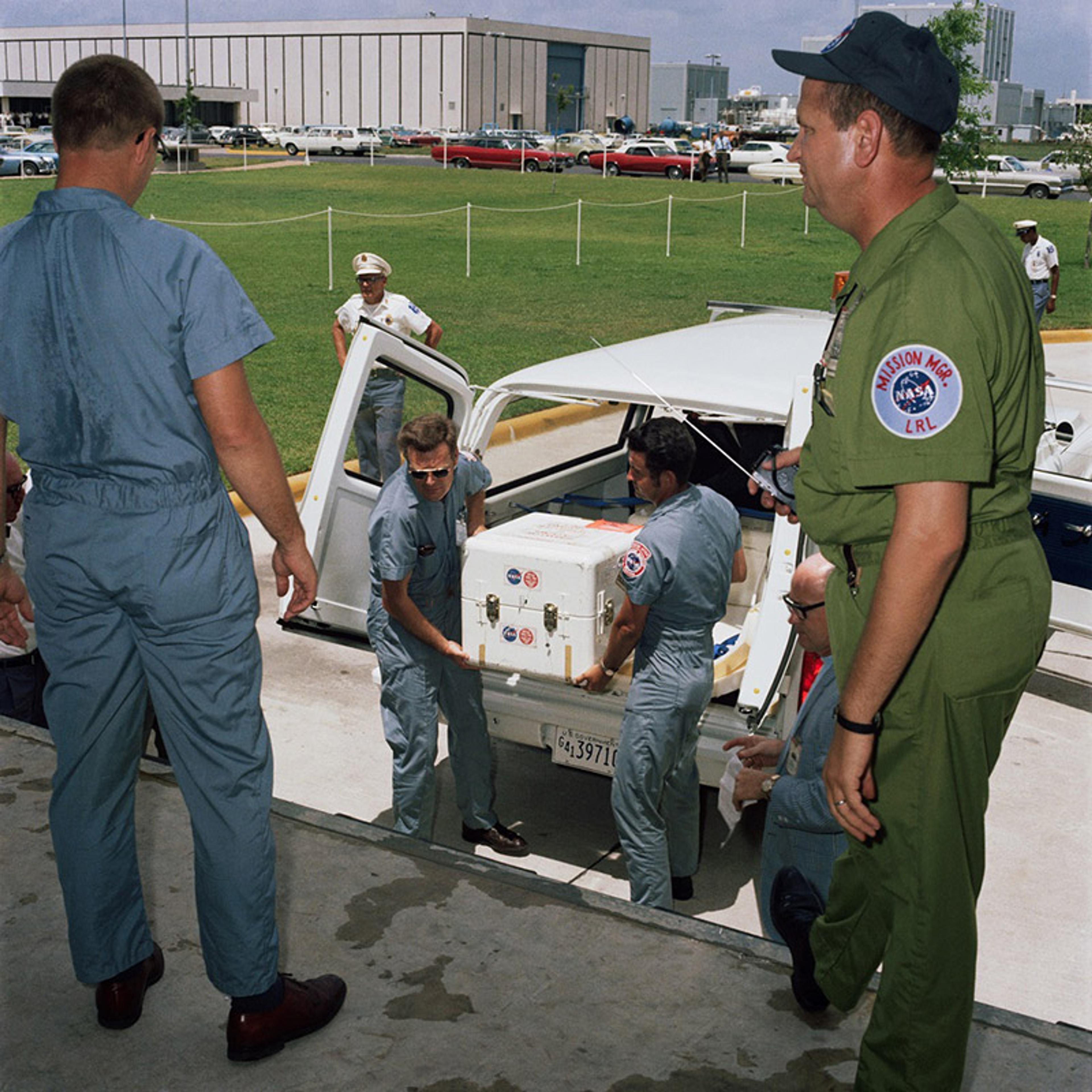 NASA personnel unloading a white container from a vehicle with a guard in the background and a man in green overalls observing.