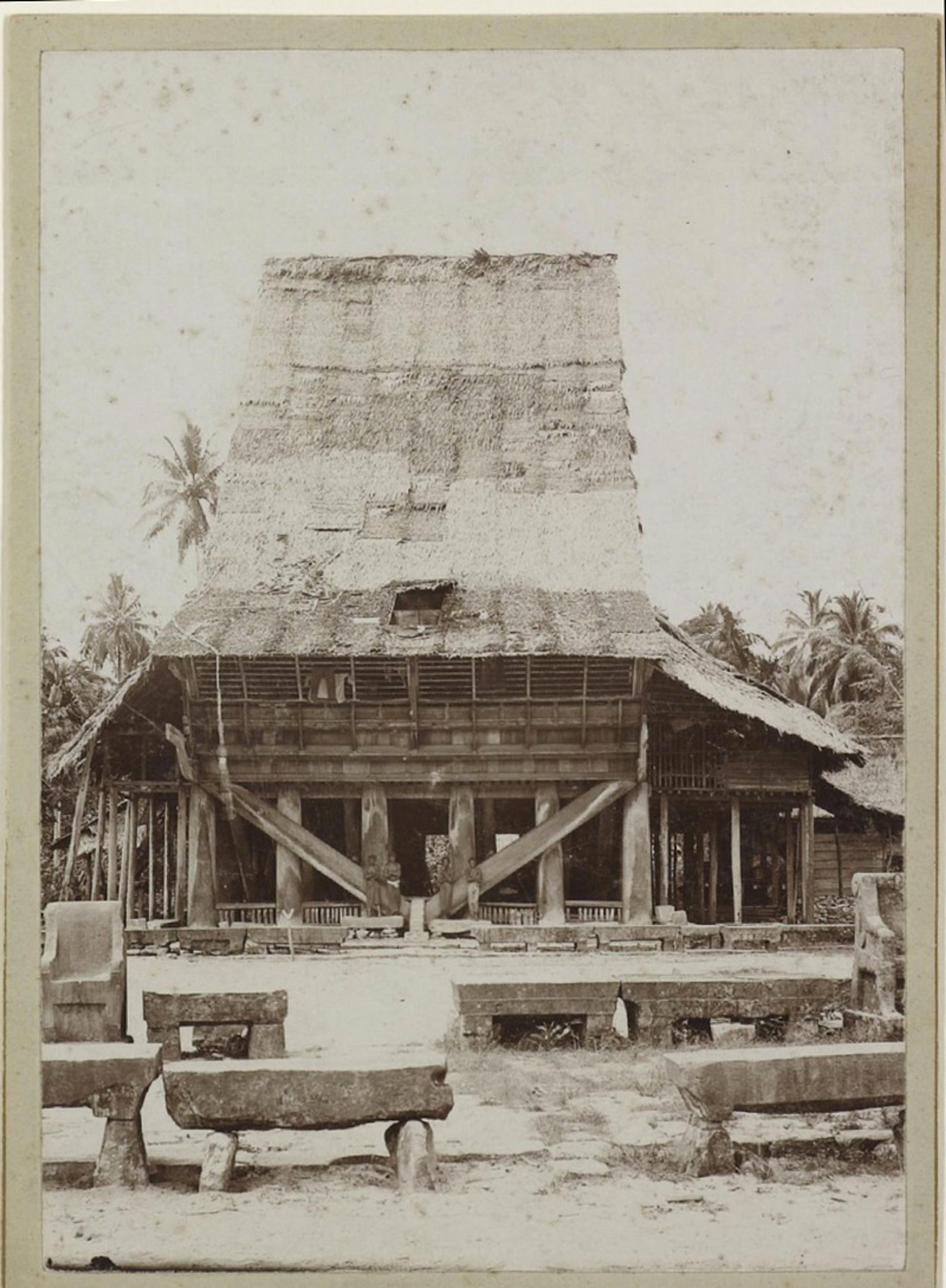 Vintage photo of a large traditional wooden house with a steep thatched roof, supported by wooden pillars, surrounded by benches.