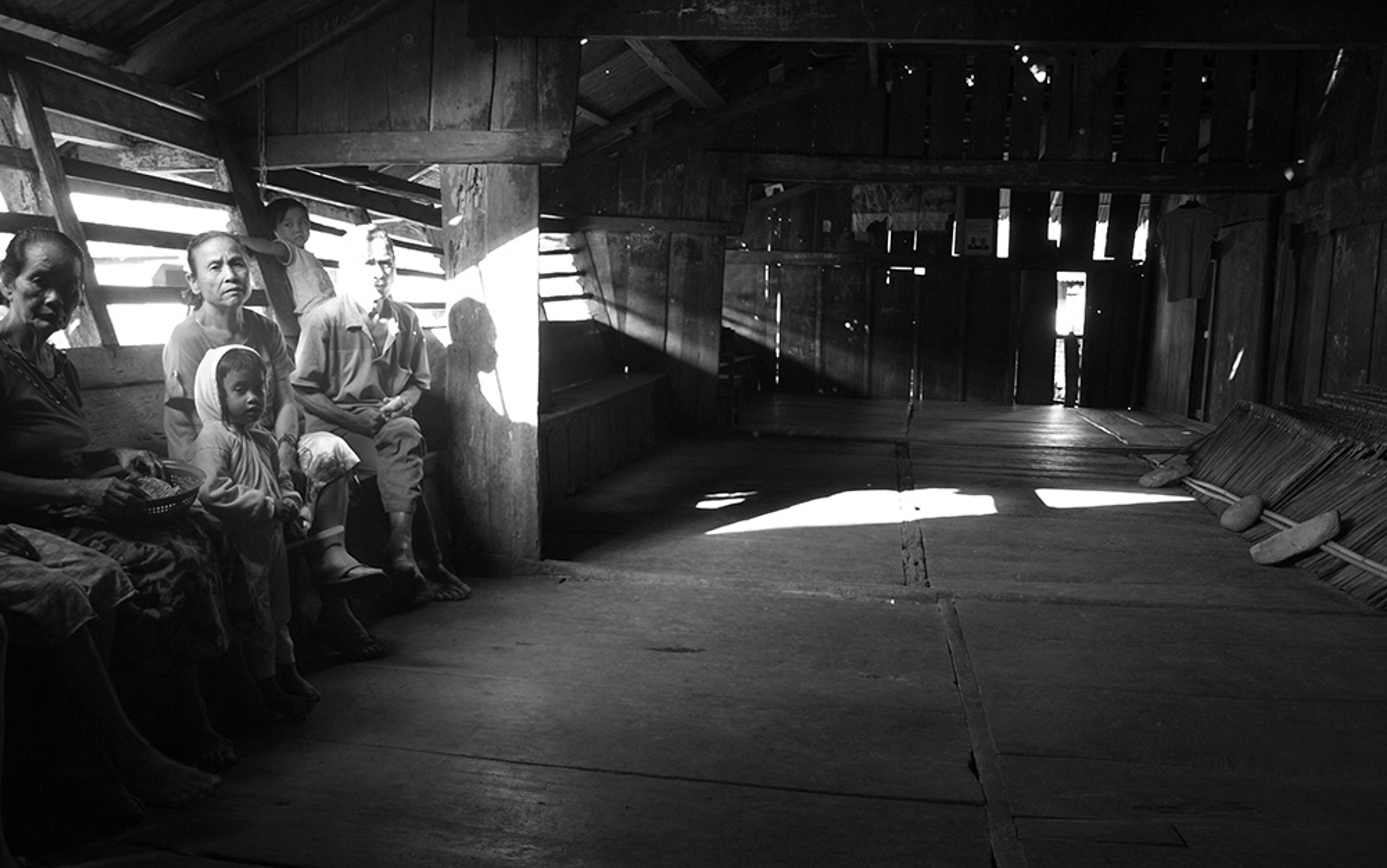 Vintage photo of people sitting on a bench in a large, dimly lit wooden room with sunlight streaming through slats.