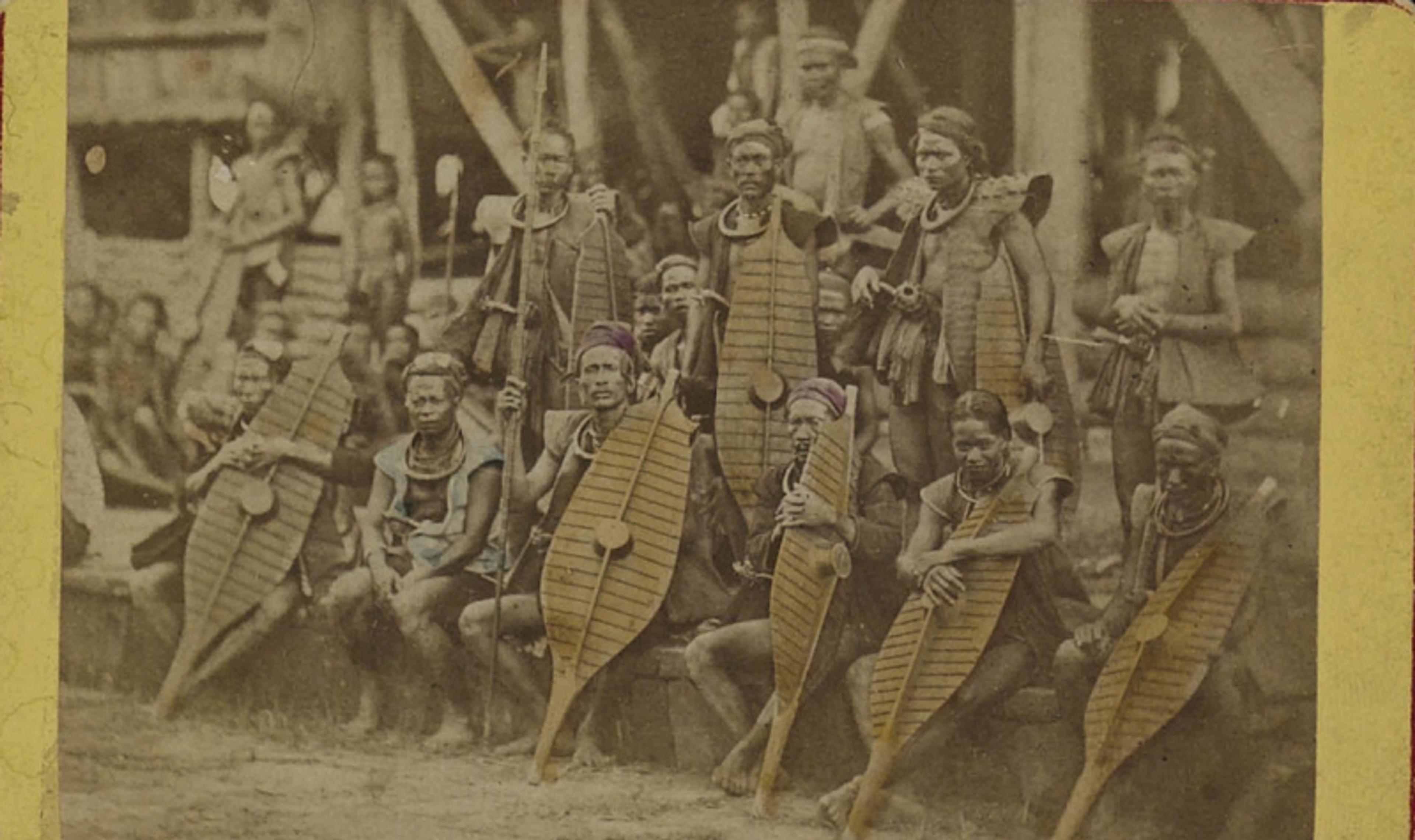 Vintage photograph of a group of people wearing traditional attire and holding shields and staffs, posed in front of a building