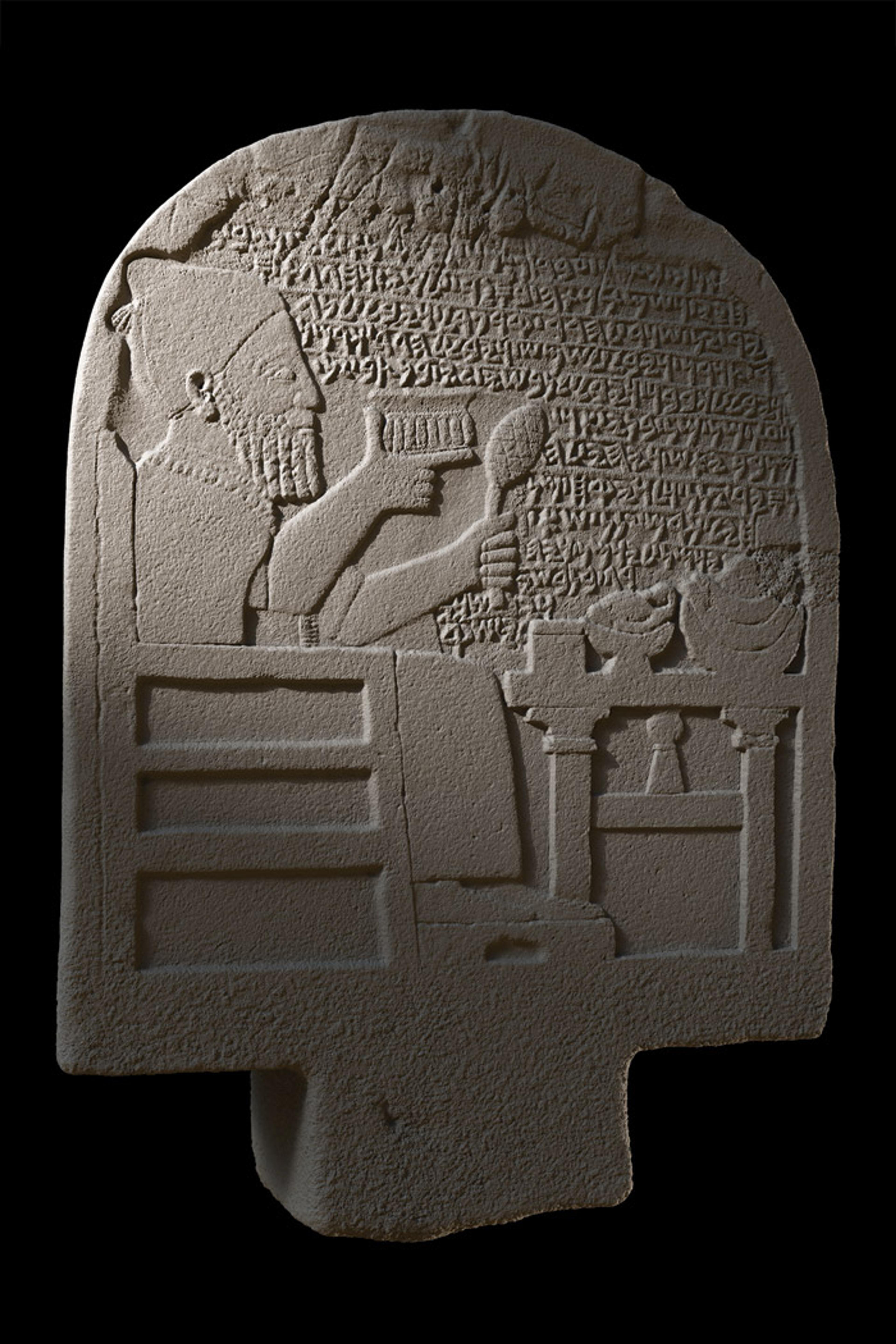 Ancient stone relief carving of a seated figure with cuneiform inscriptions, holding a goblet and spoon, objects on a table nearby.