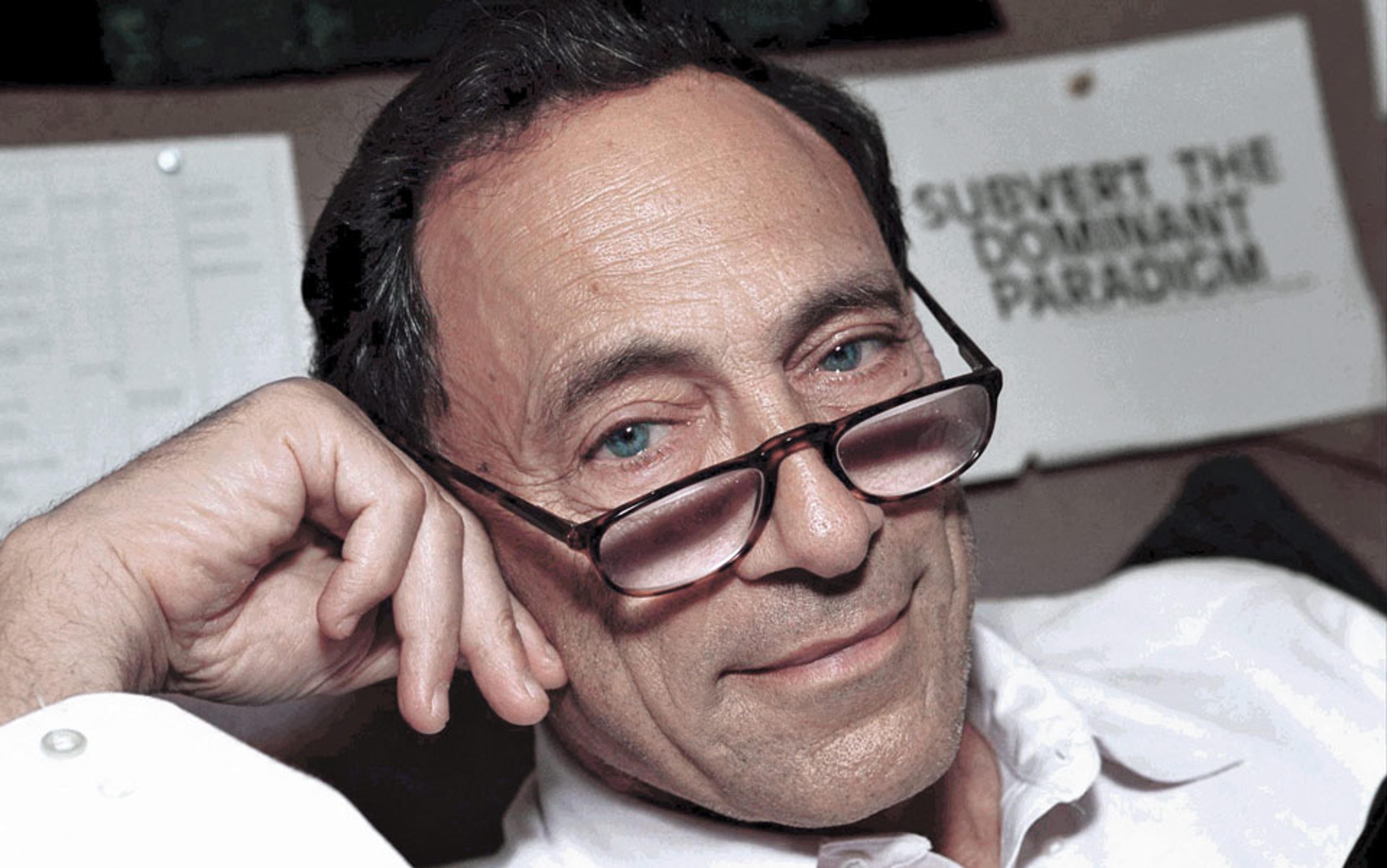 A man with glasses, wearing a white shirt, resting his head on his hand in front of a sign reading “Subvert the dominant paradigm.”