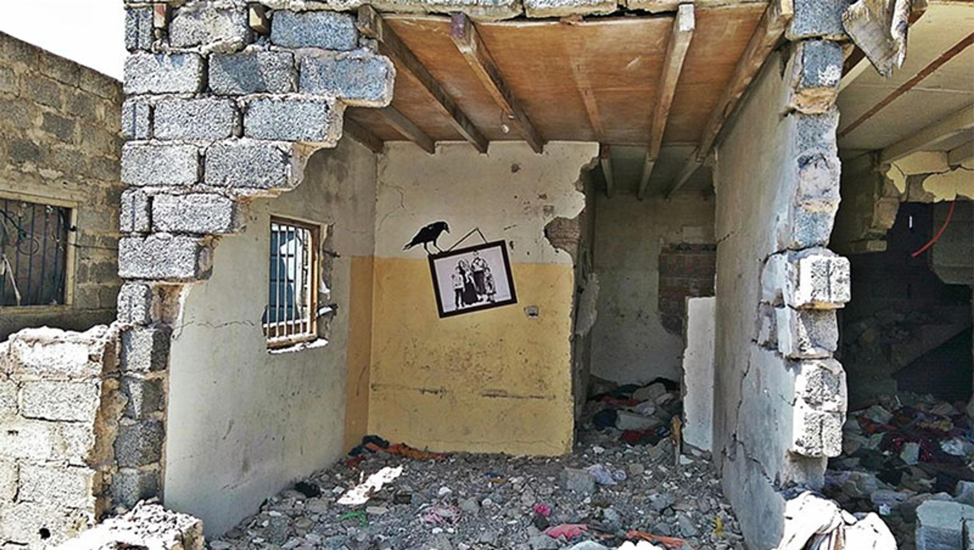 A damaged building interior with rubble on the floor, missing walls, and a mural of a framed family picture hanging askew with a crow on top.