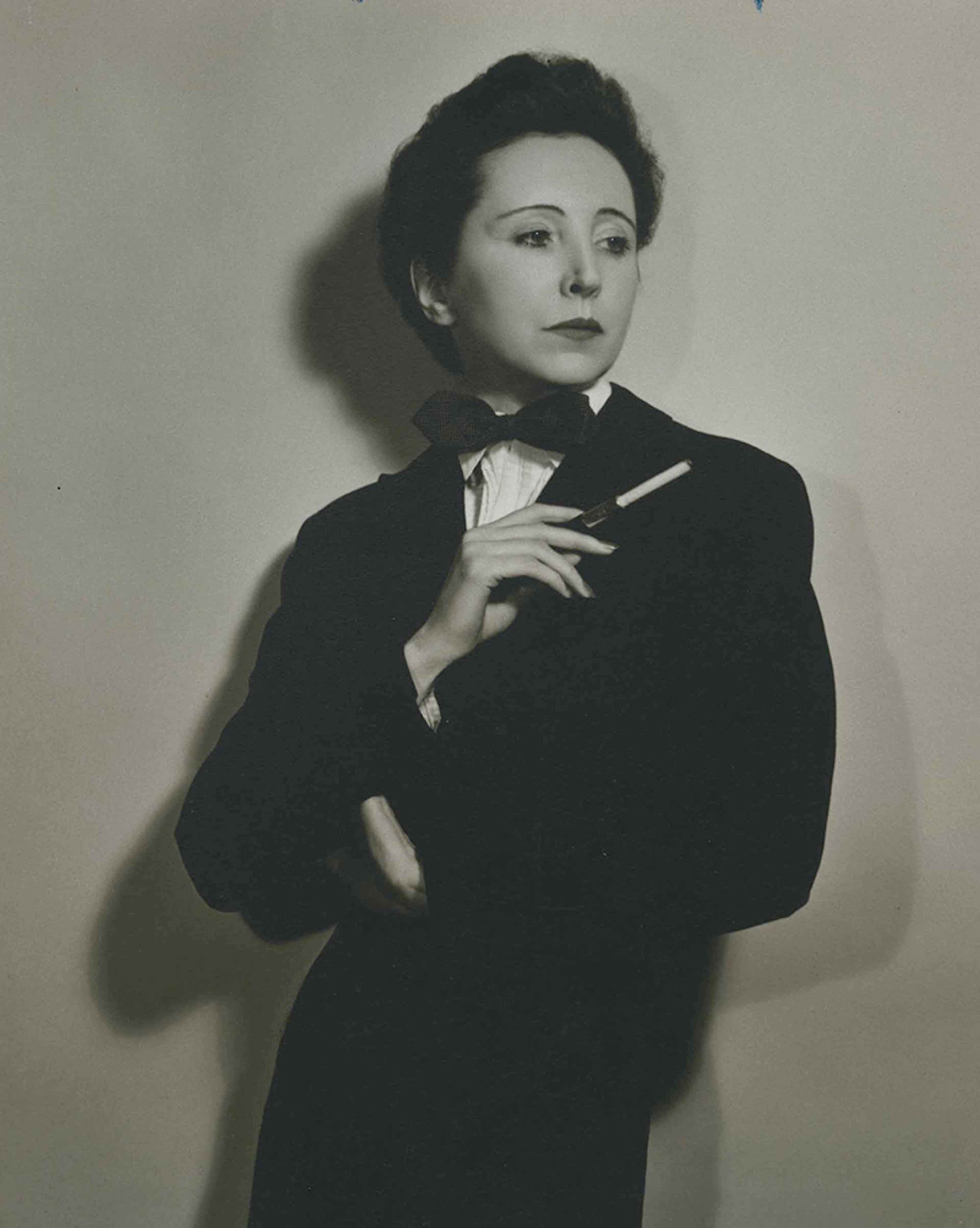 Vintage photo of a woman in a suit, bow tie and white shirt holding a cigarette in a holder with a thoughtful expression.