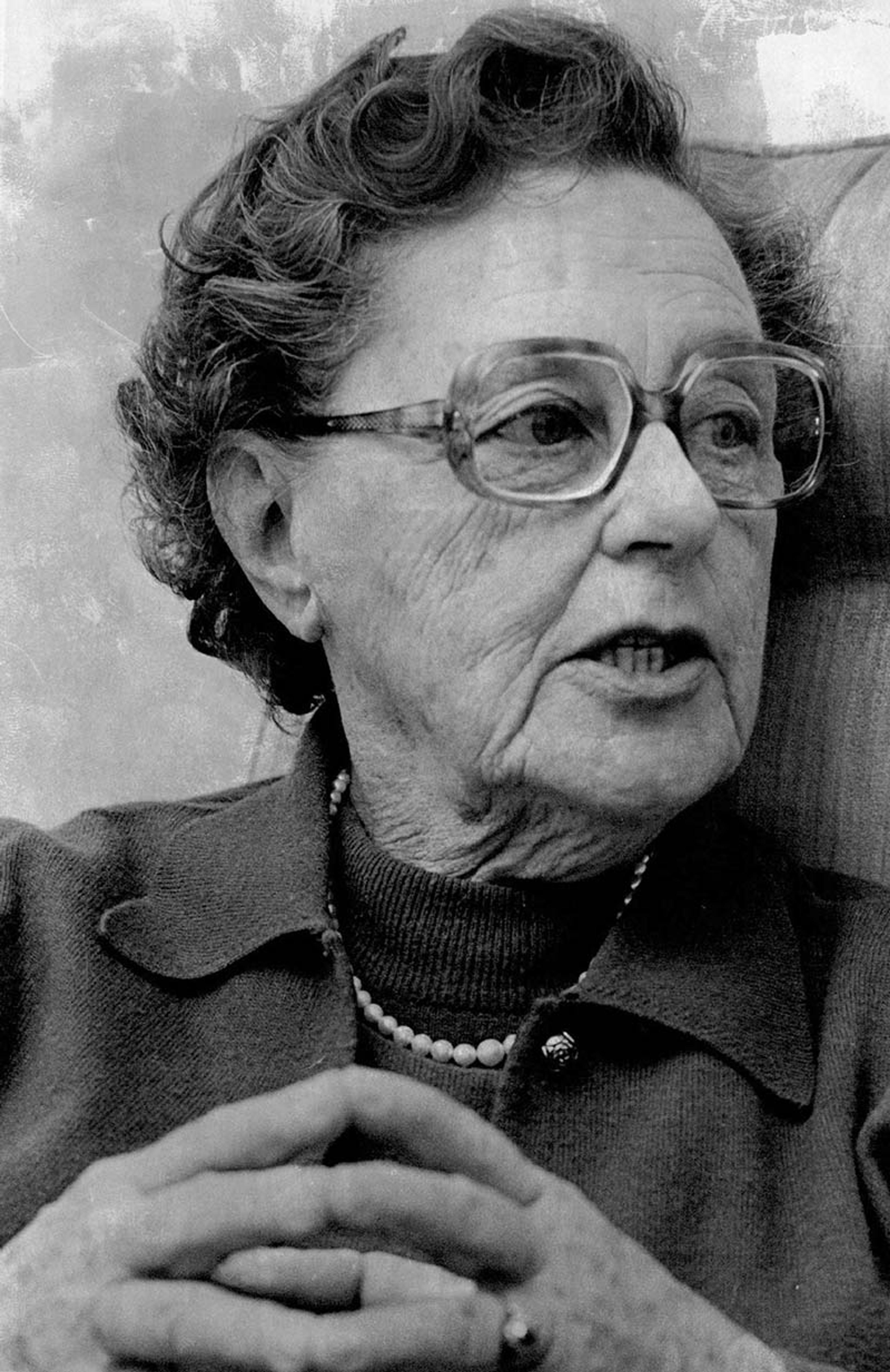 Vintage photo of an elderly woman with glasses, wearing a collared shirt and necklace, speaking with her hands clasped.
