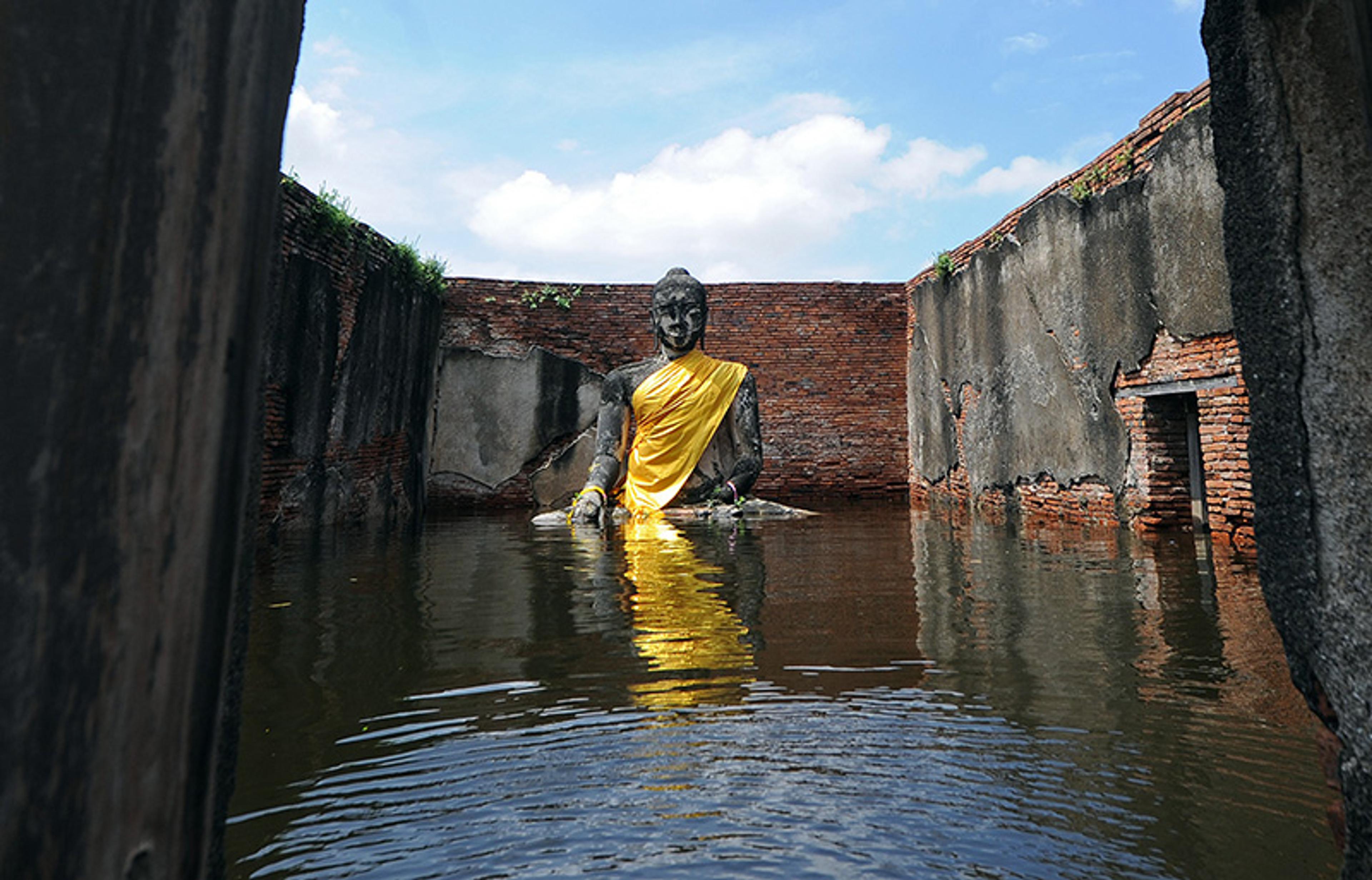 Photo of a large Buddha statue wrapped in a yellow robe, partially submerged in water within the ruins of a brick-walled structure.