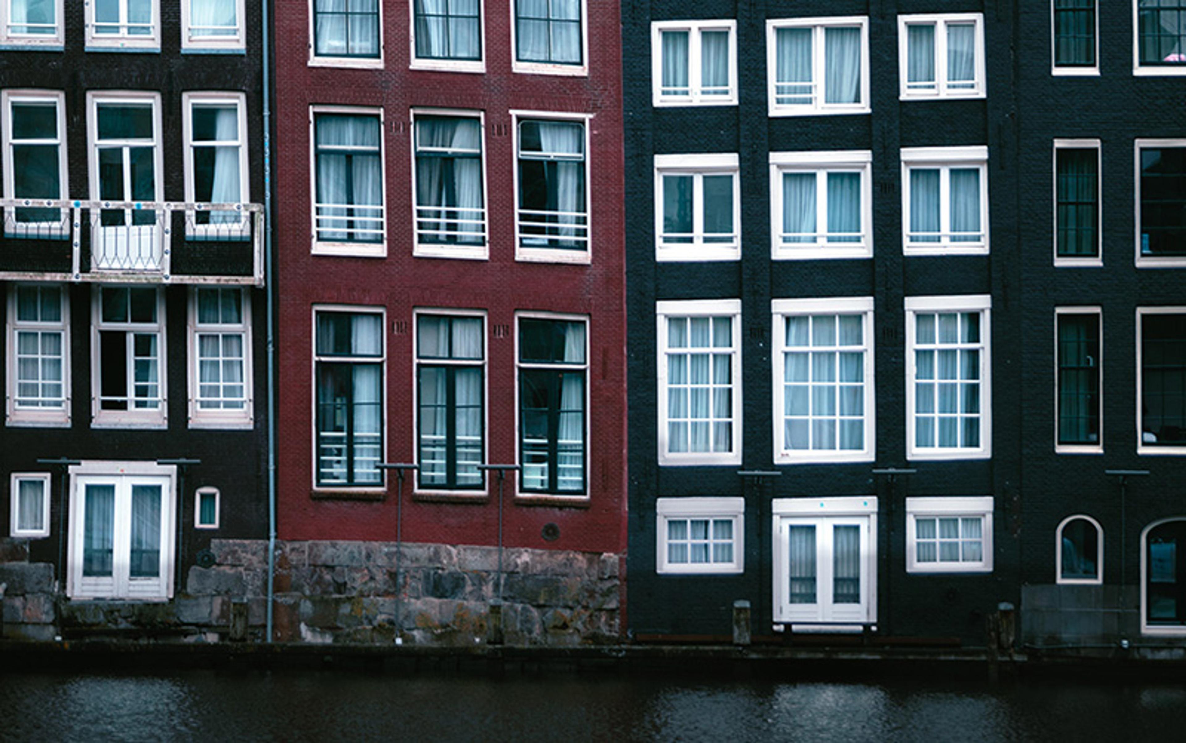 Photo of three adjacent canal houses with large windows in varied dark colours and a red central building, reflecting in water.