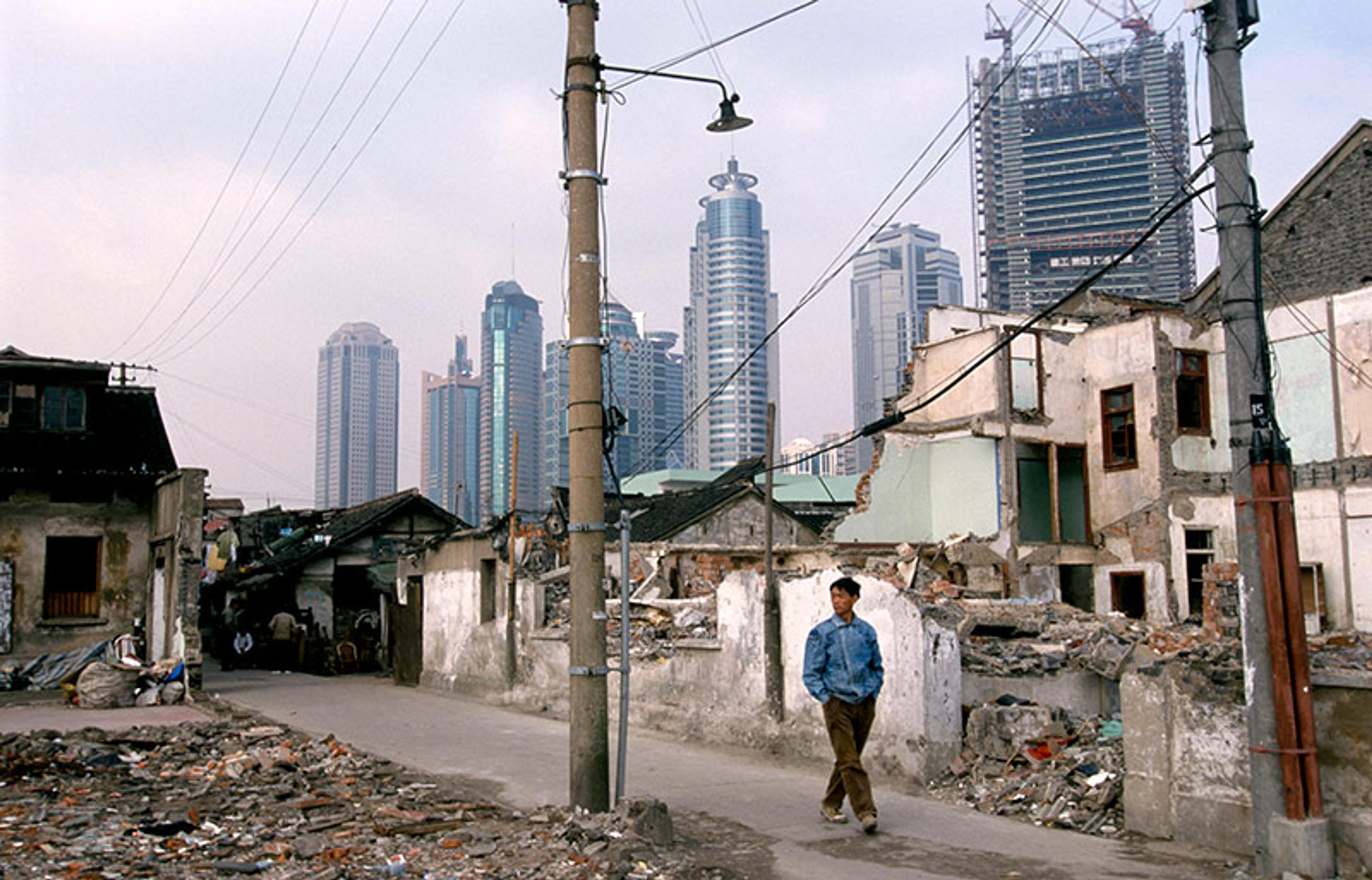 A man walking past dilapidated buildings in an urban area with modern skyscrapers in the background, under a cloudy sky.