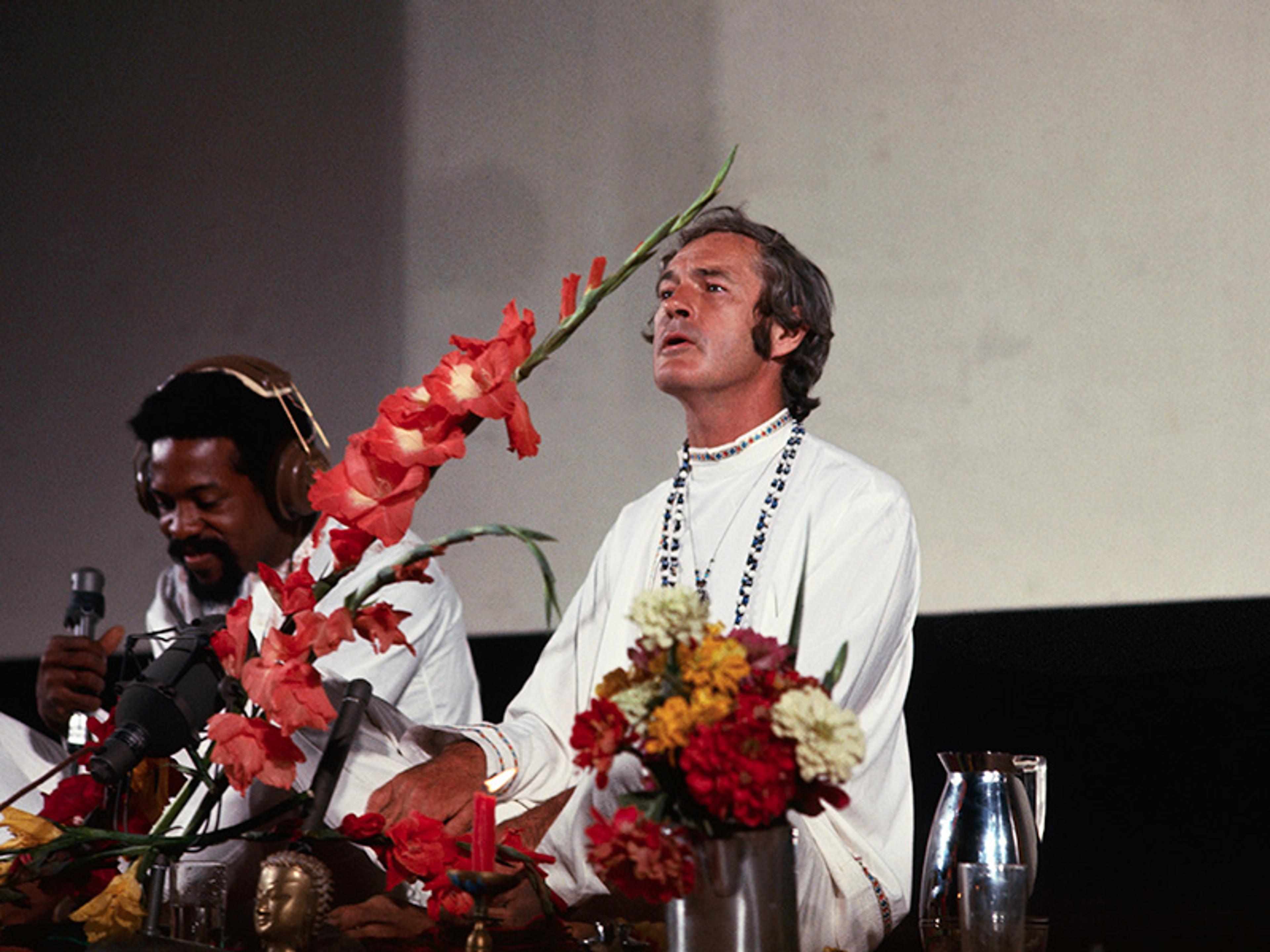 A man wearing white and beads, holding flowers, accompanied by another man with headphones speaking into a microphone.