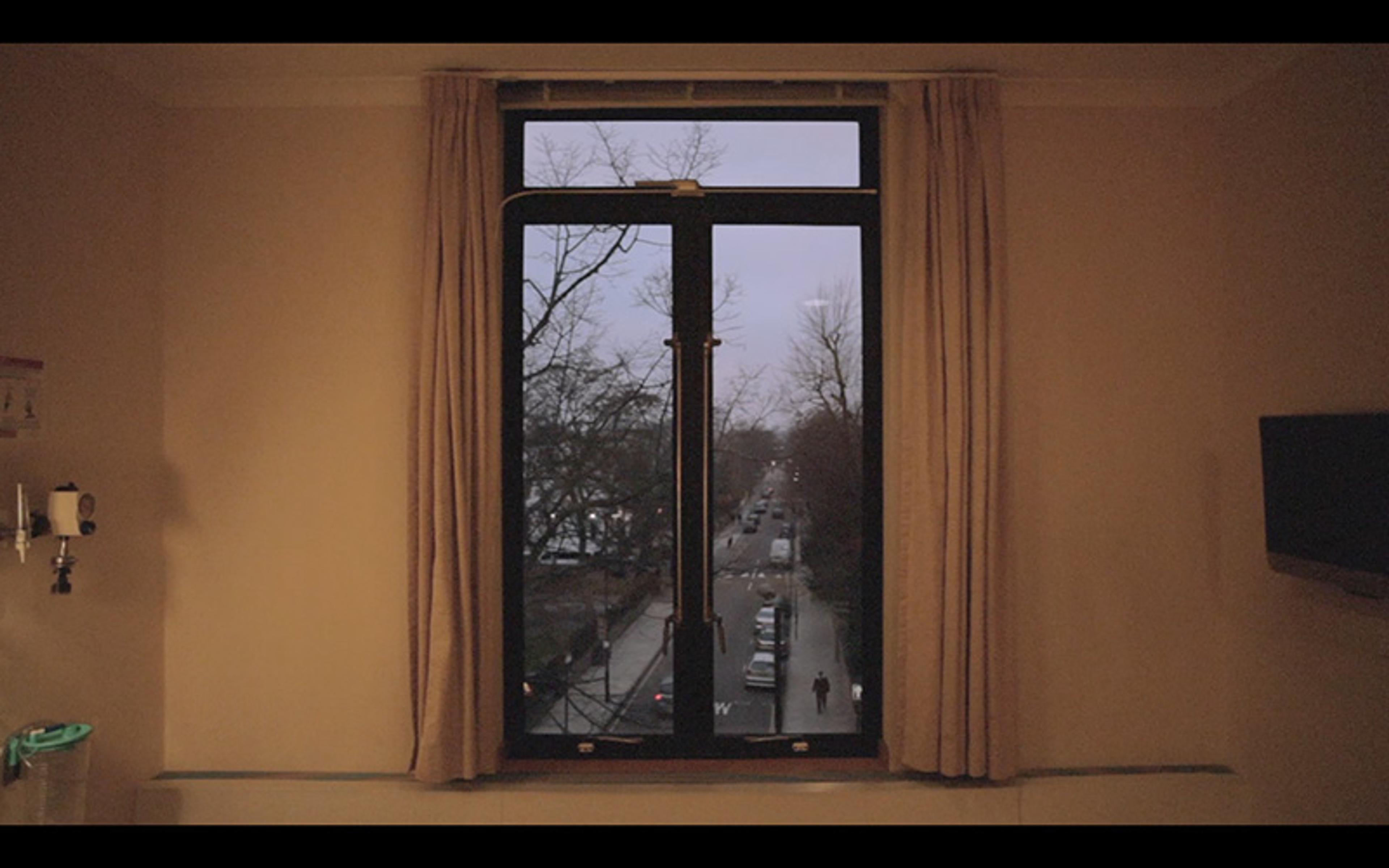 Photo of a room with a window showing a street lined with trees outside. Curtains frame the window, and a TV is on the right wall.
