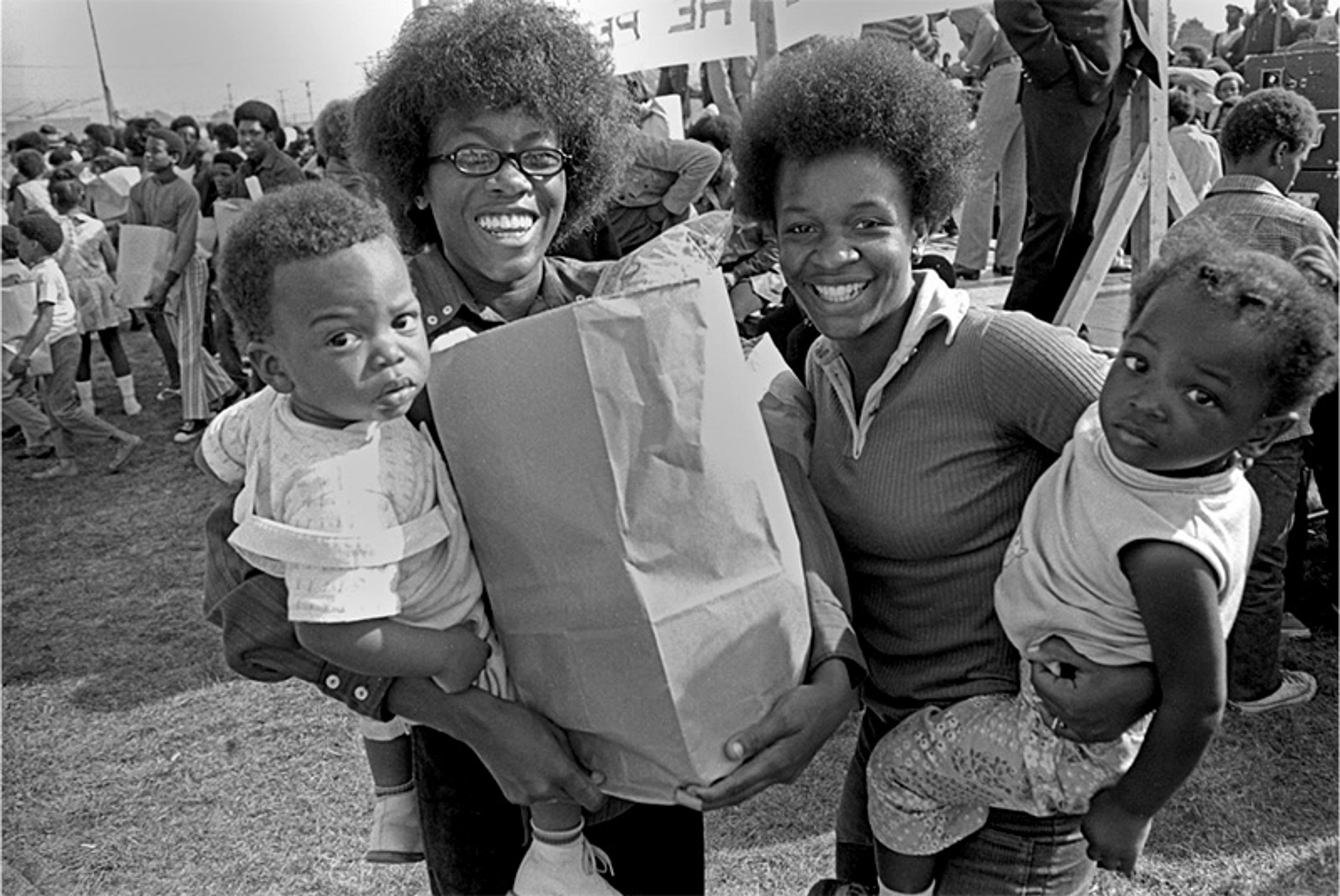 Black and white photo of two women holding children and a paper bag, smiling at a community event with a crowd in the background.