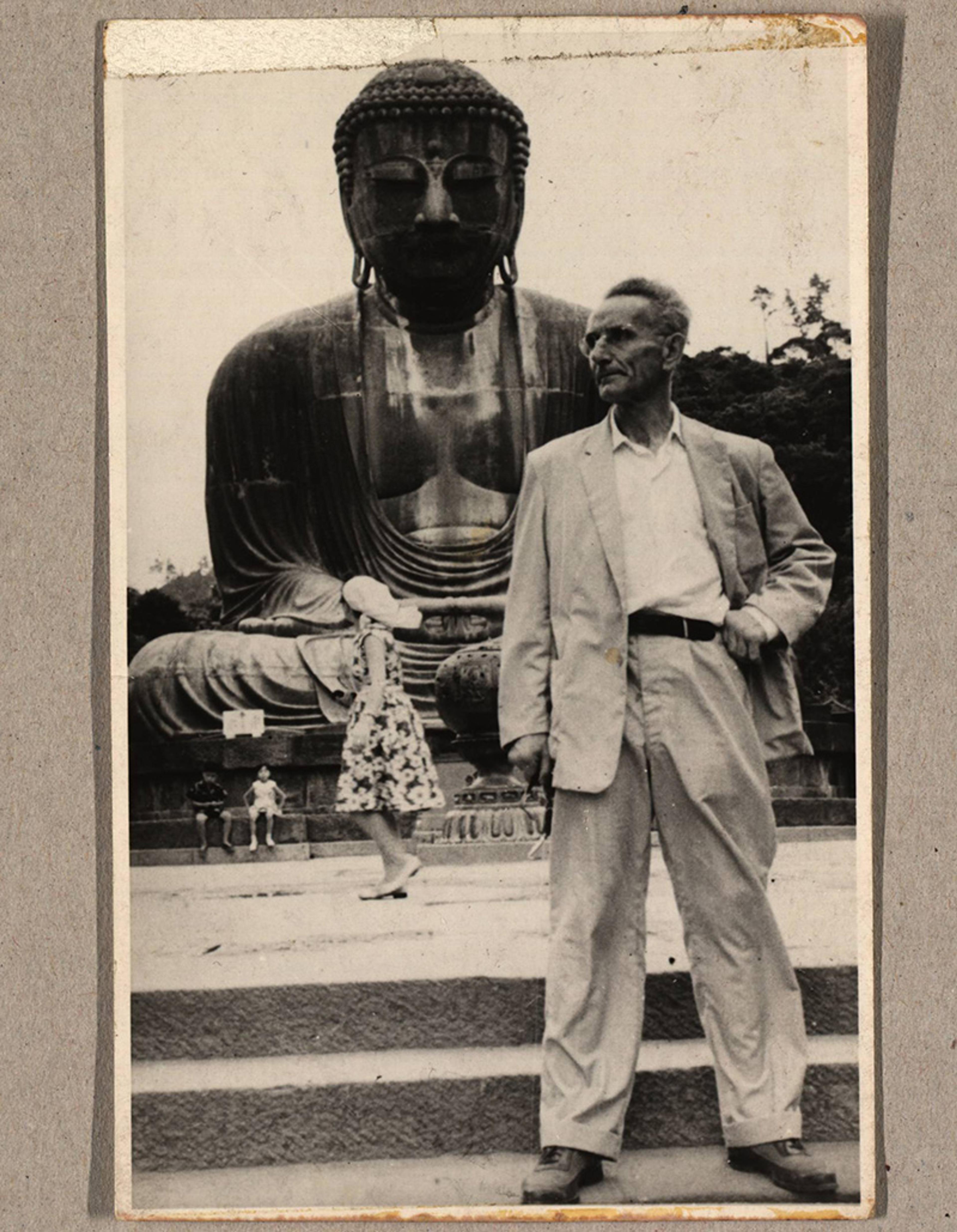 Black-and-white photo of a man in a suit standing in front of a large seated Buddha statue with trees in the background.