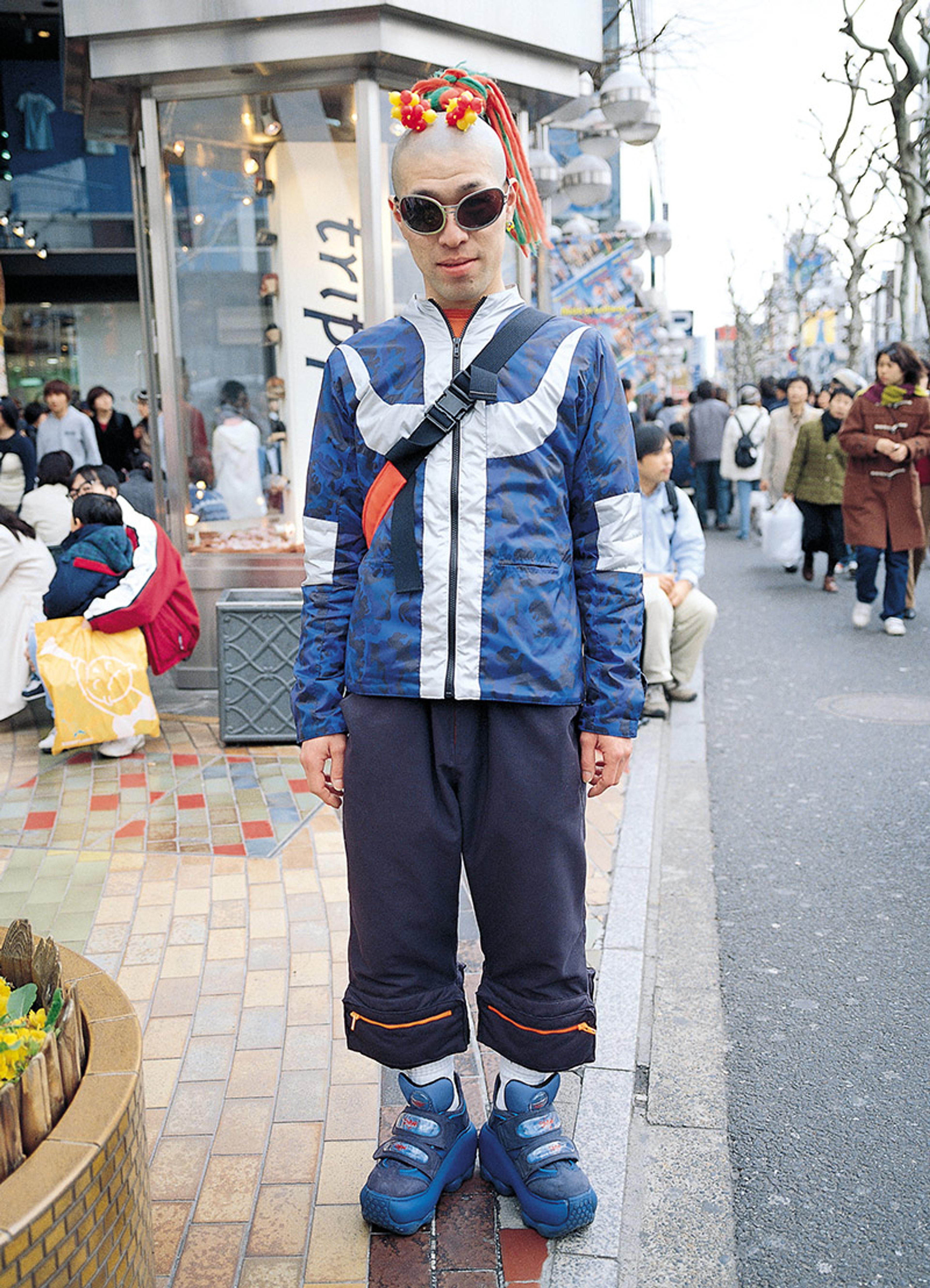 A Japanese man in a blue jacket, sunglasses and colourful hair on a busy urban street. People and shops are visible in the background.
