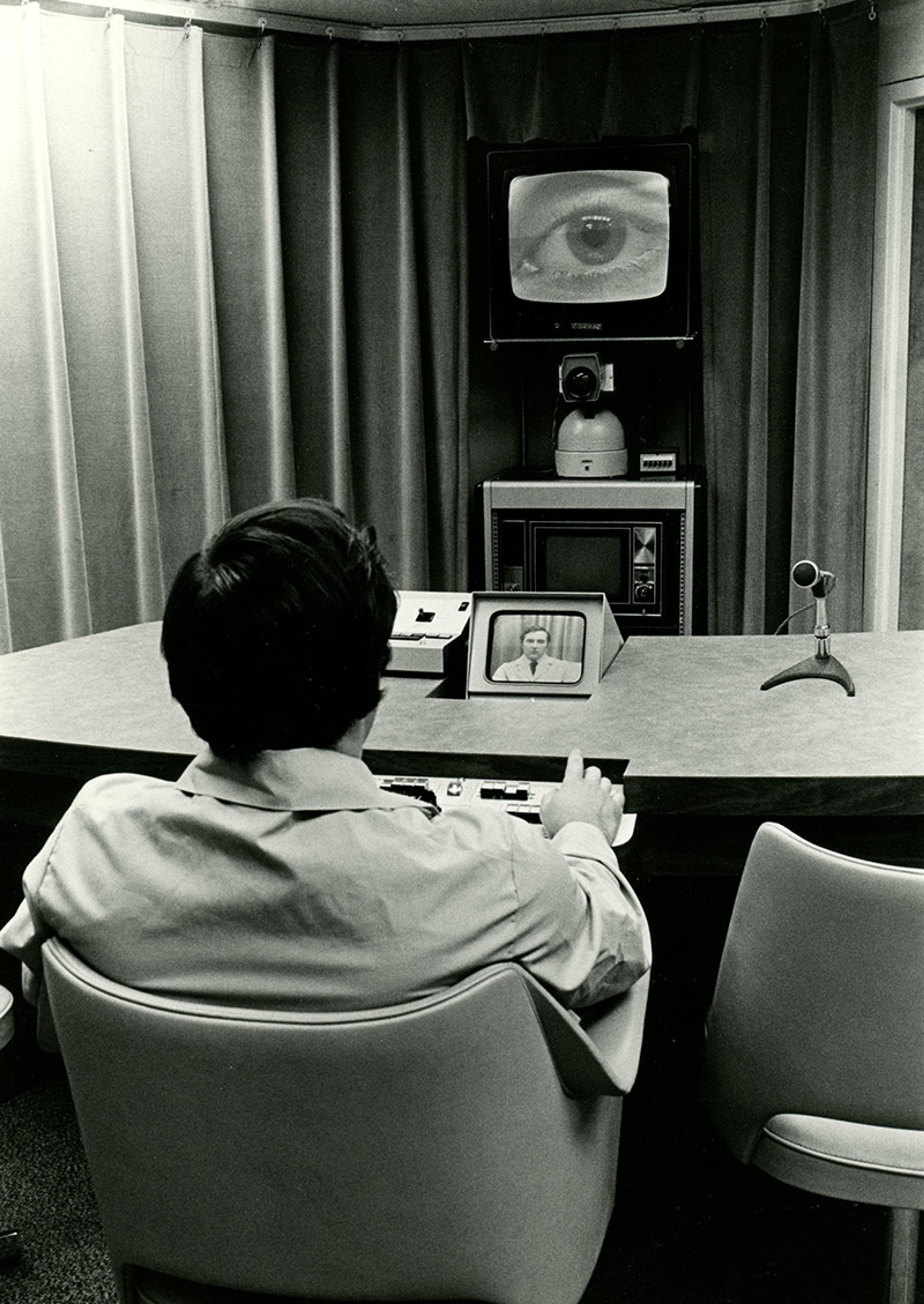 Vintage photo of a man in front of consoles, a small monitor showing a person, and a large screen displaying an eye.