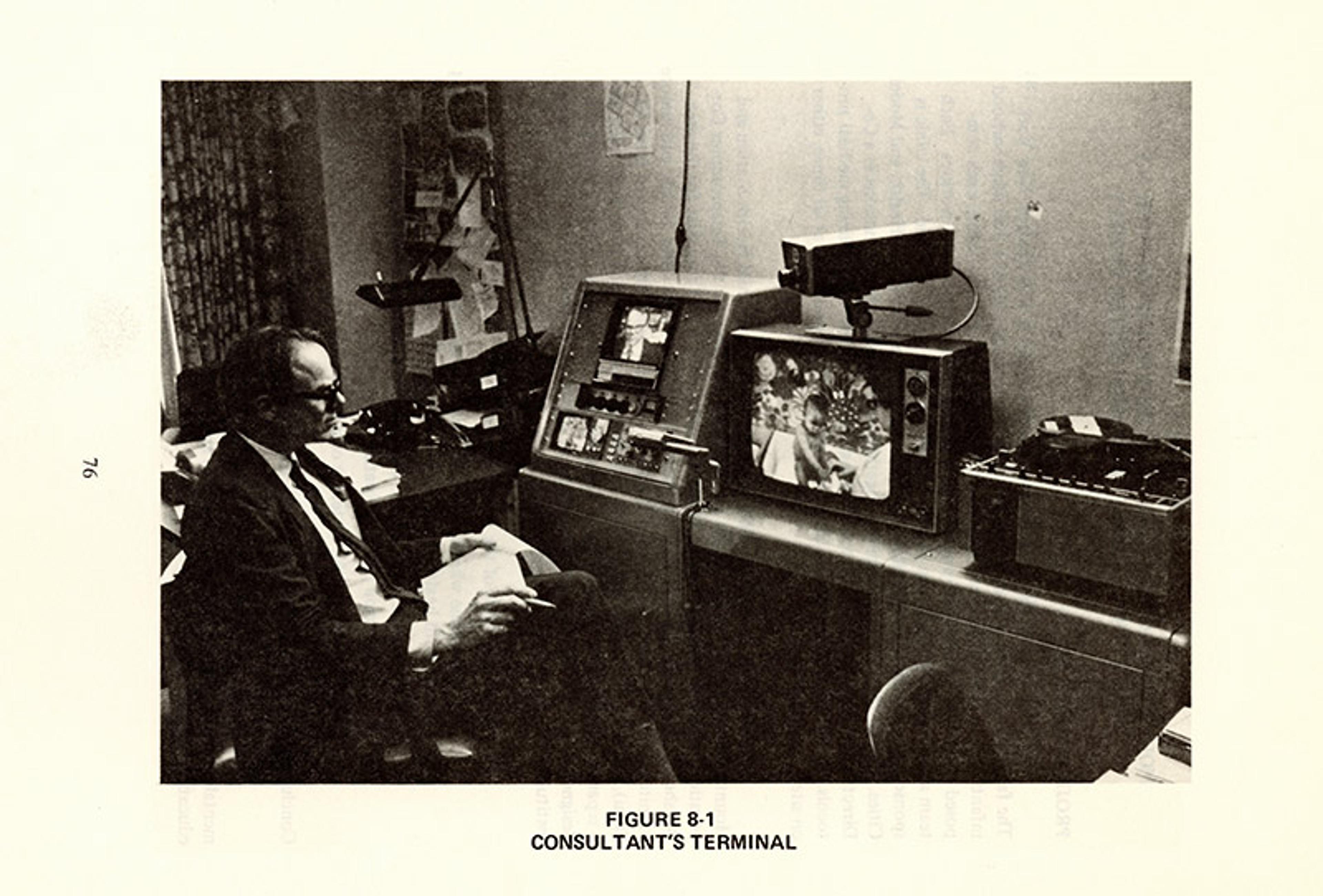Vintage photo of a man seated at a desk with a computer terminal and monitor displaying an image in an office setting.