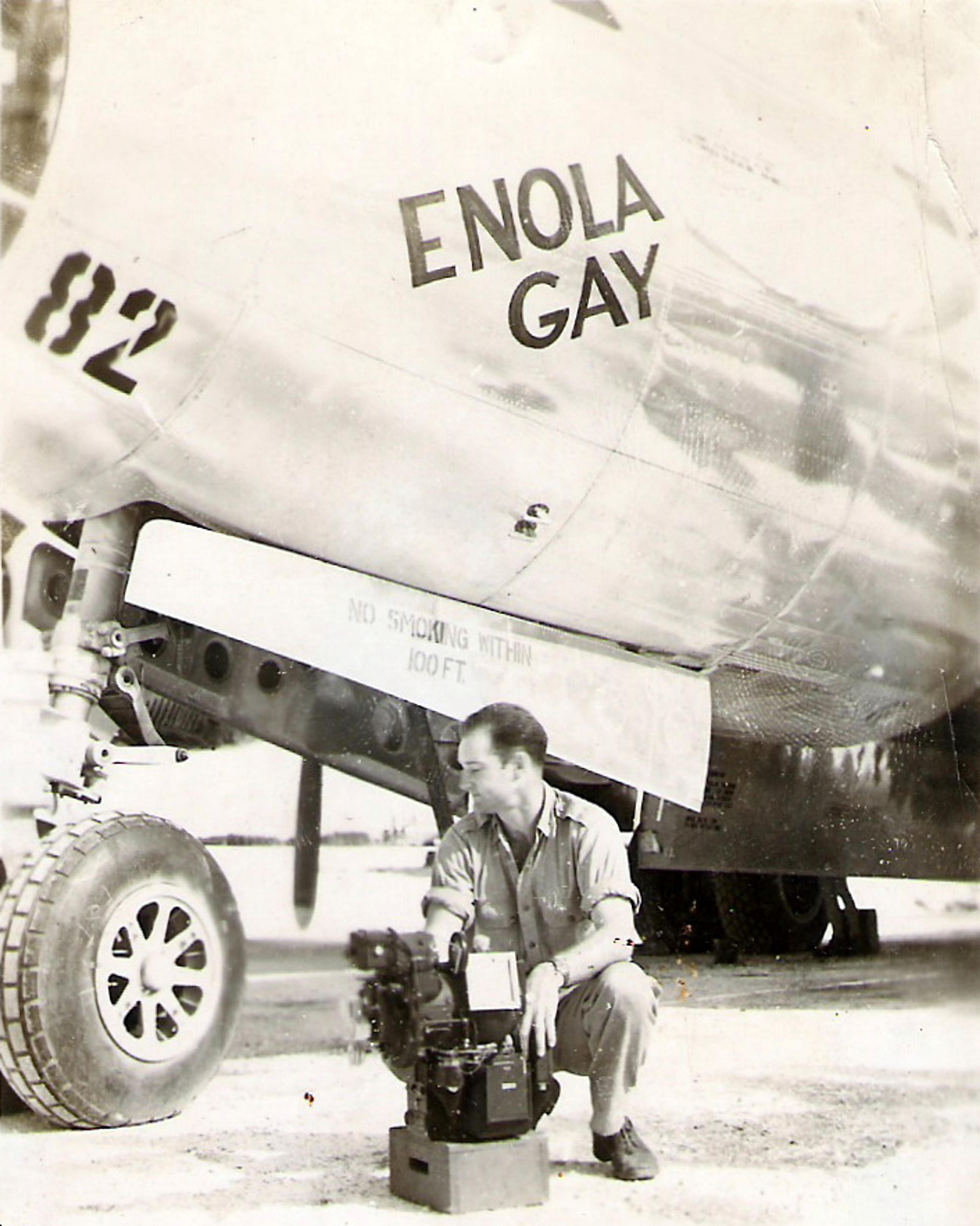 Vintage photo of a man crouching beside a “Enola Gay” plane with equipment. The plane’s nose and landing gear are visible.