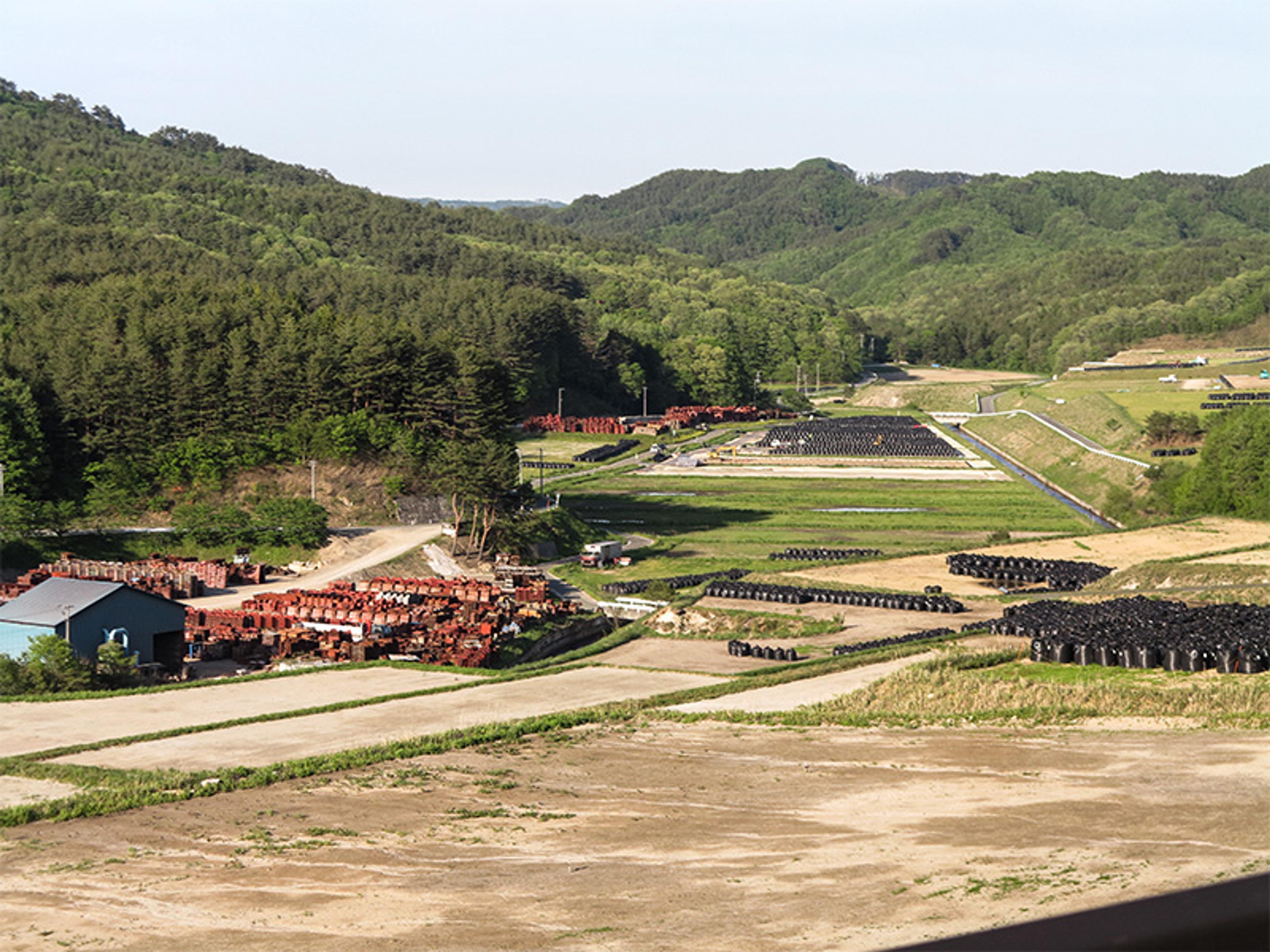Aerial photo of a rural landscape with stacks of barrels, green hills in the background, and a road running through the area.
