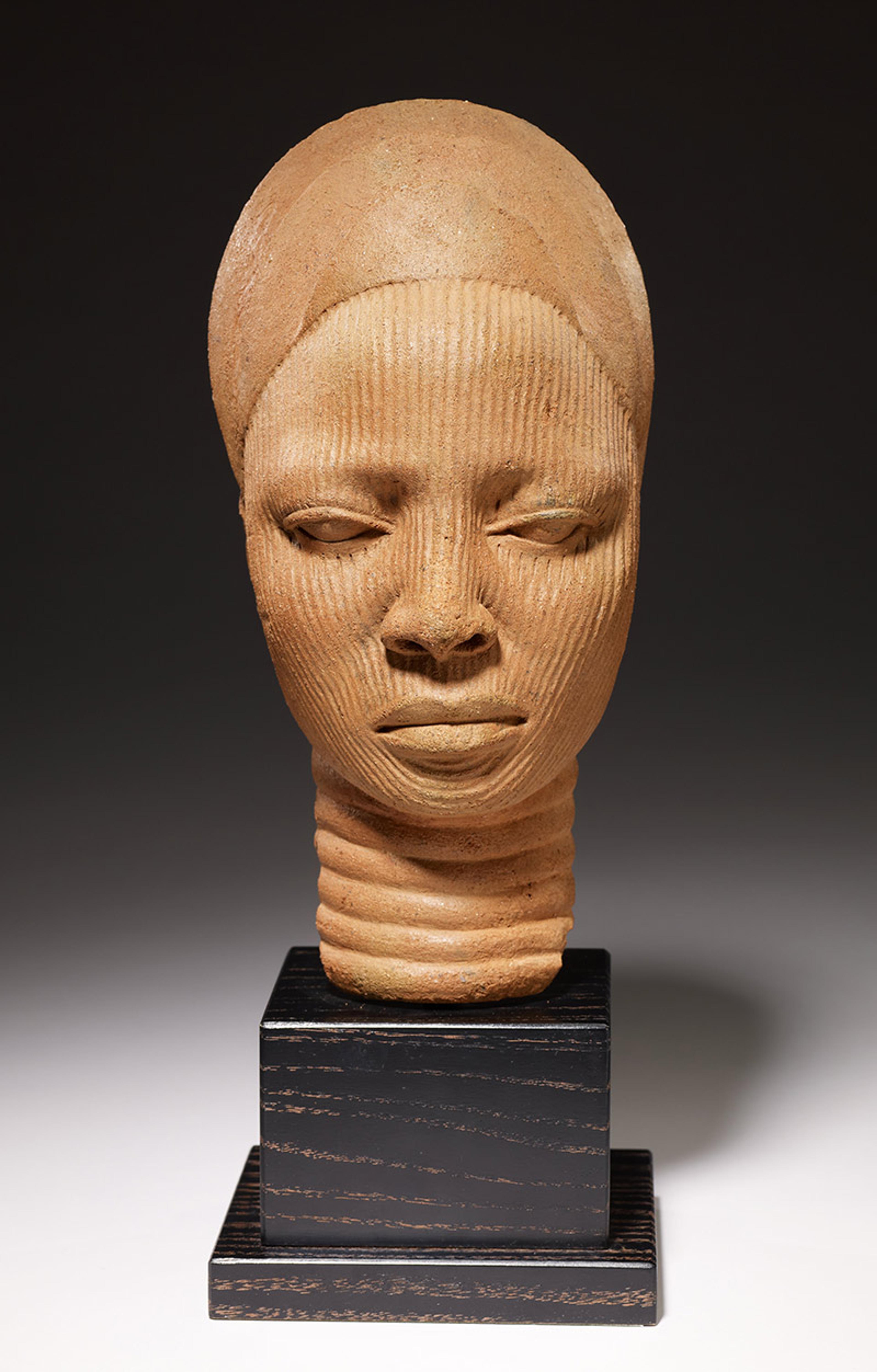 Terracotta sculpture of a head with detailed features, mounted on a black pedestal, displayed against a dark background.