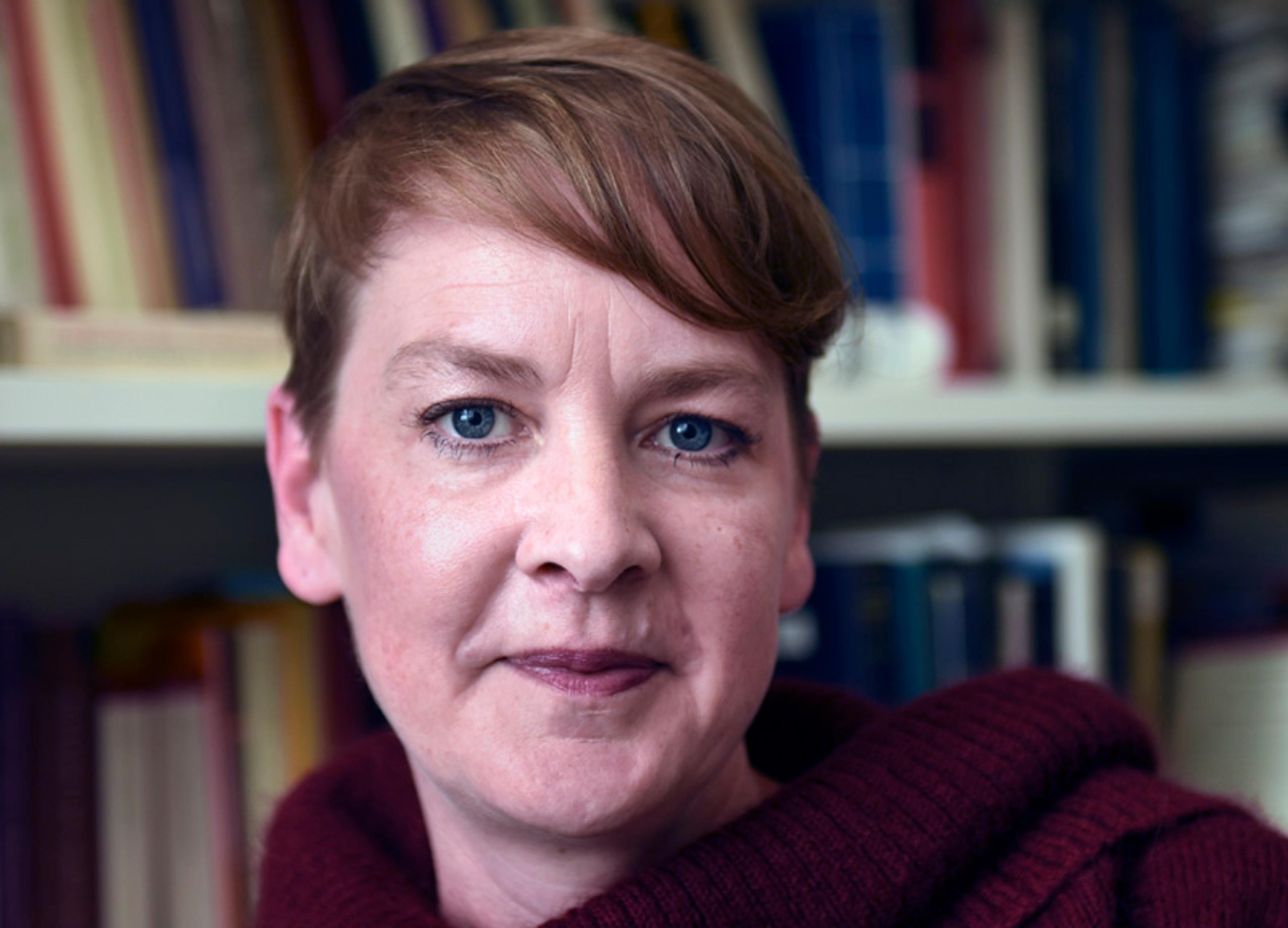 A woman with short hair and blue eyes wearing a maroon sweater, standing in front of a bookshelf filled with books.