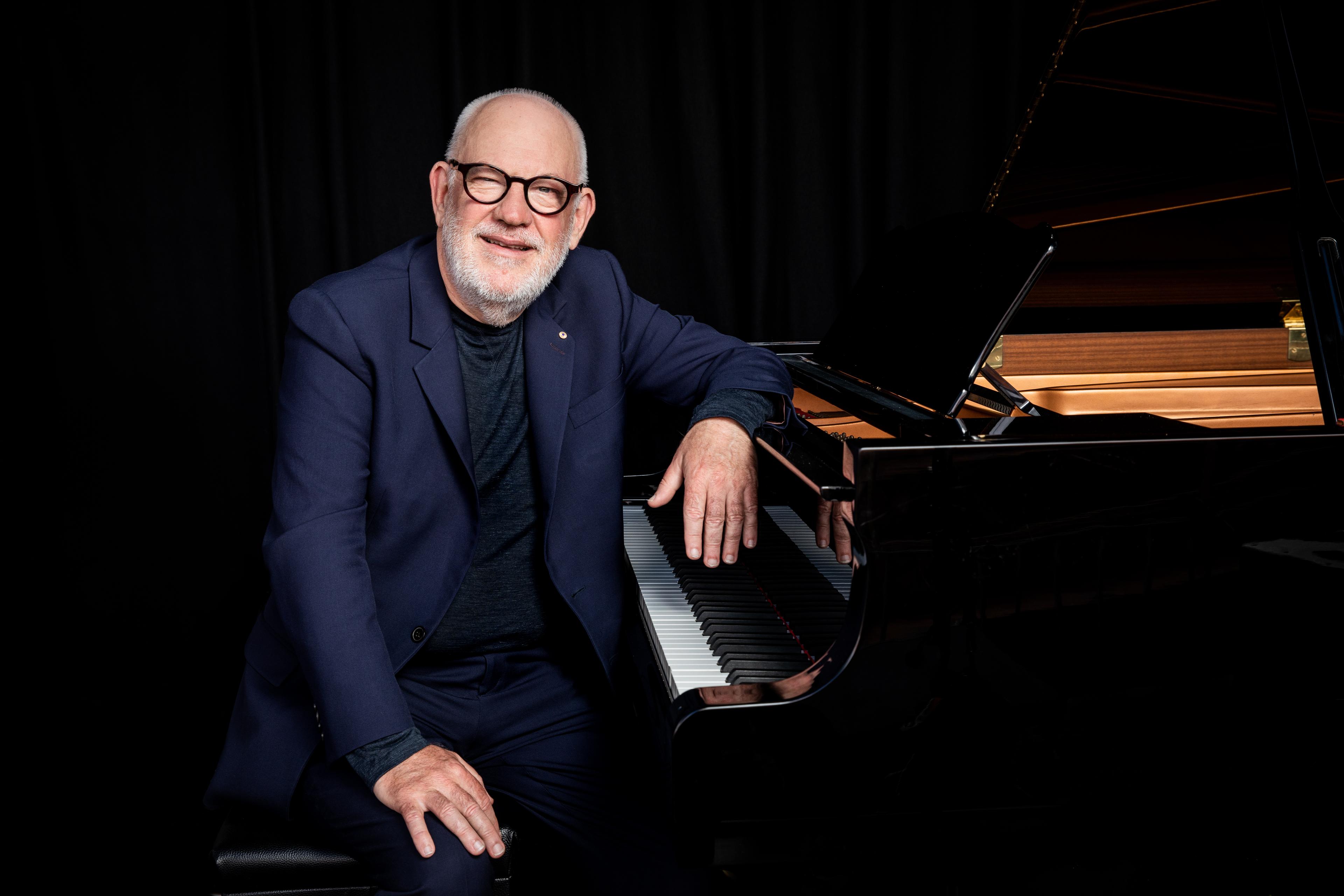 An elderly man with glasses and a beard, dressed in a suit, seated at a grand piano, smiling against a dark background.