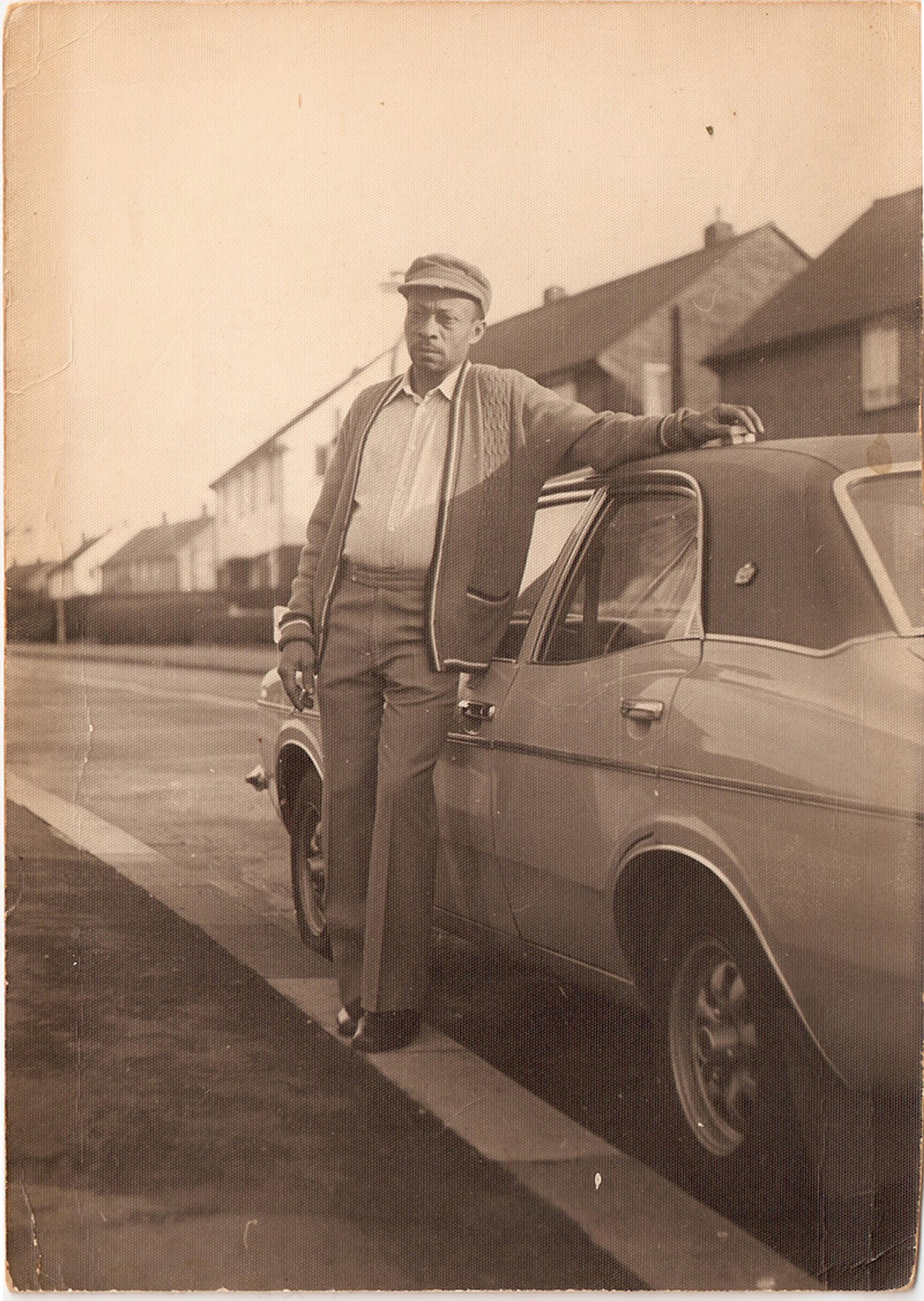 A vintage photo of a man in a cap leaning on a classic car parked on a suburban street in front of houses.