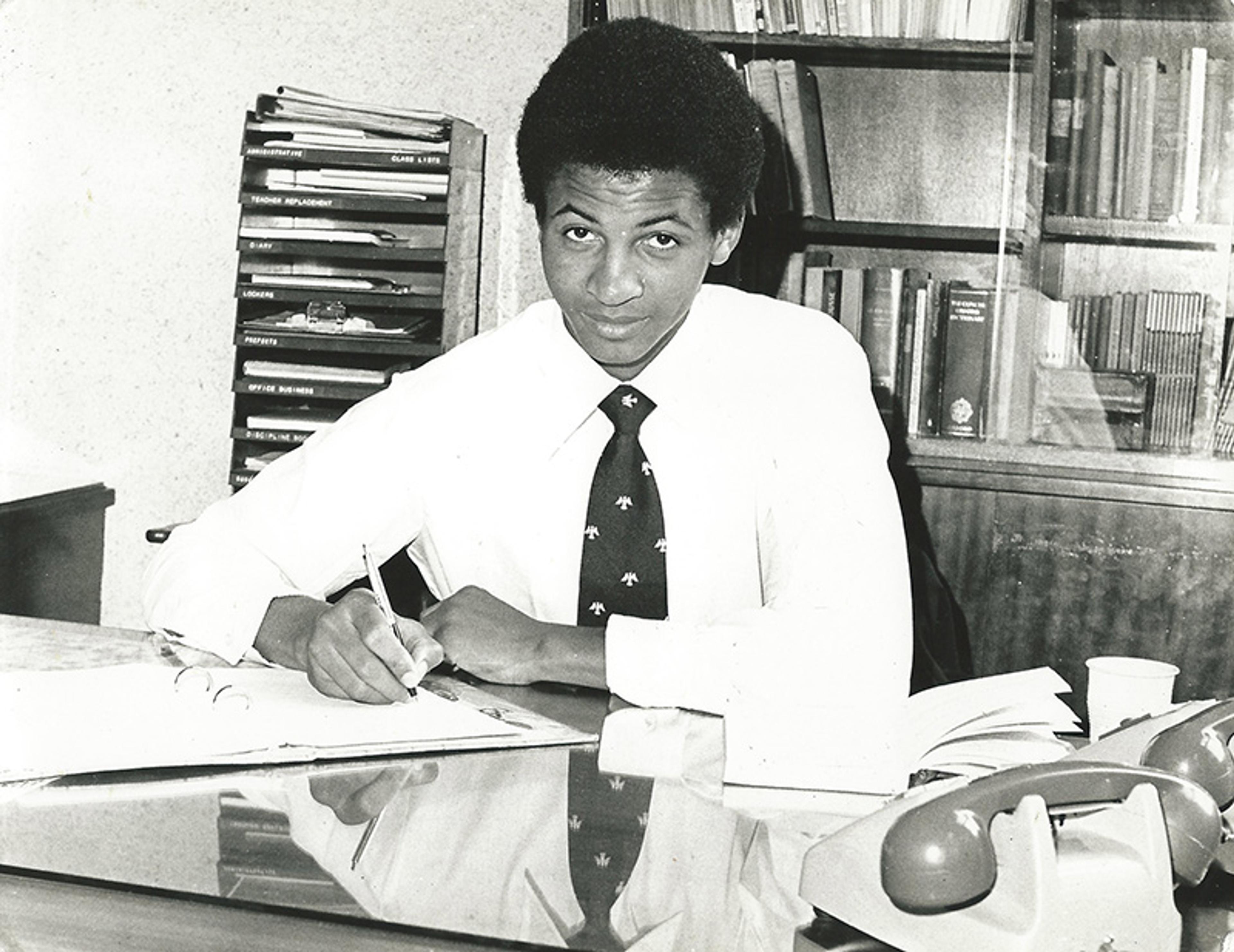 Black-and-white photo of a man in a shirt and tie, seated at a desk with books and a telephone, writing on paper.