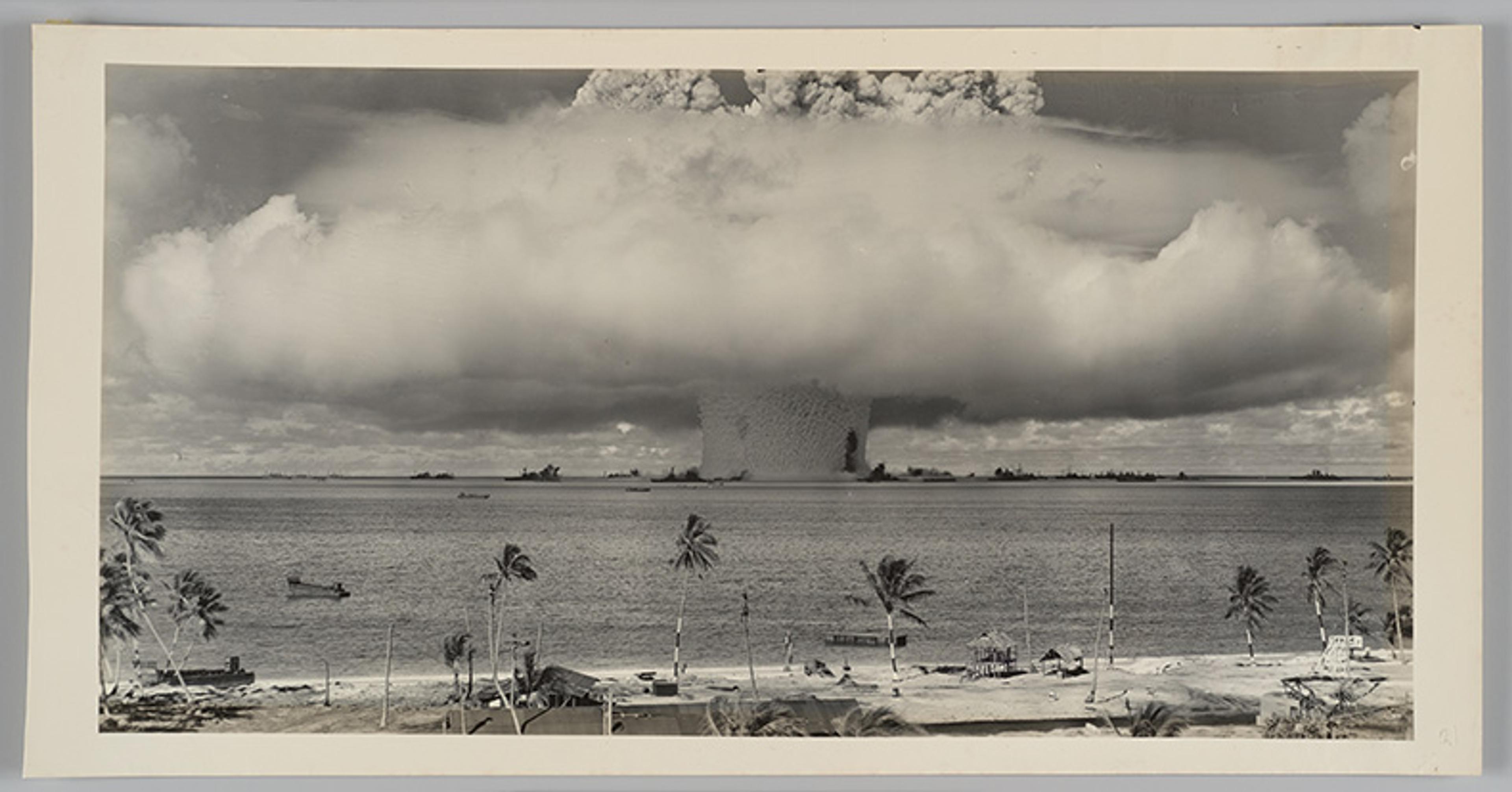 Black-and-white photo of a nuclear explosion over the ocean viewed from shore with palm trees and small structures in the foreground.