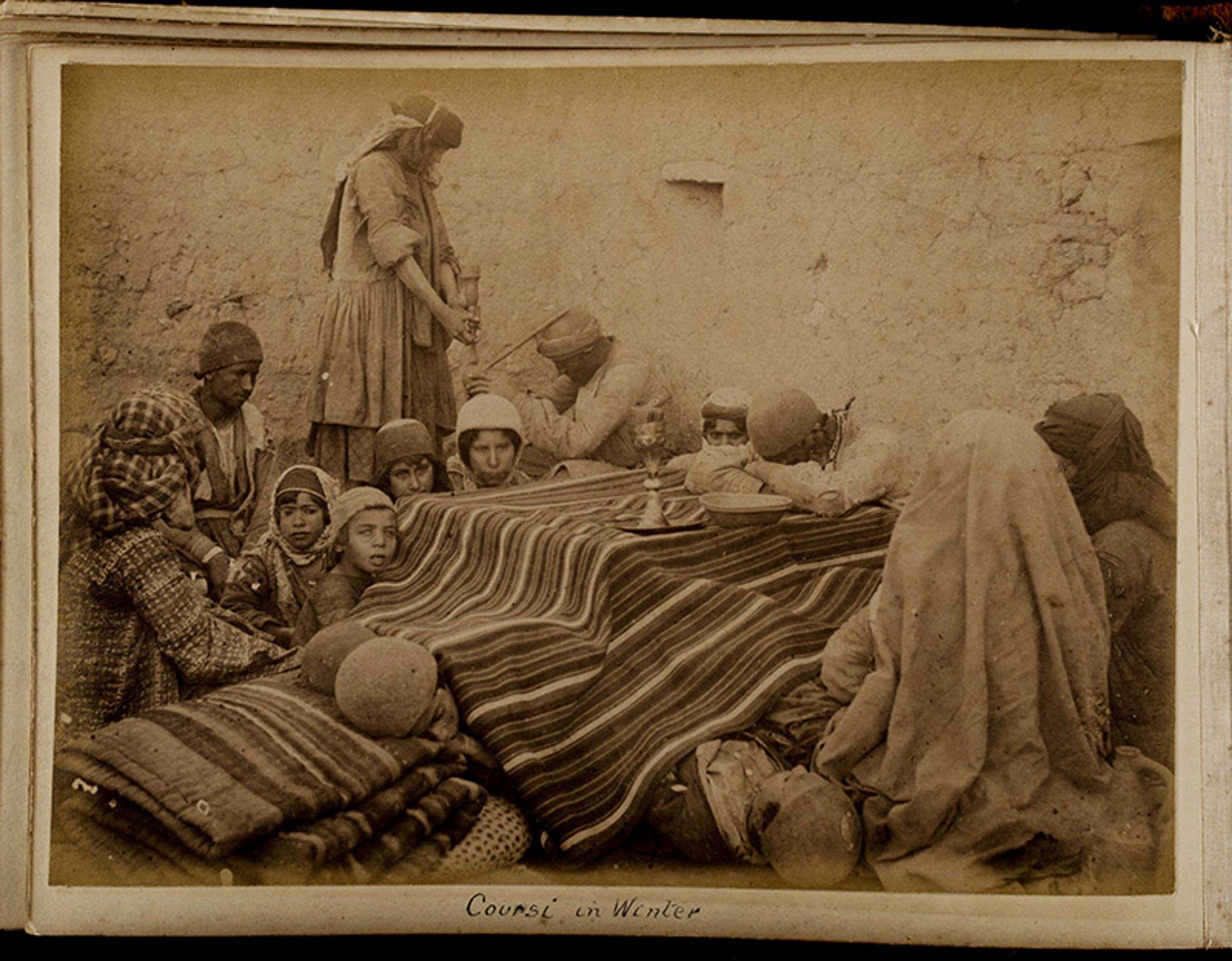 Black-and-white photo of people gathered around a table covered with a striped cloth set against a wall, labelled “Coursi in Winter”.