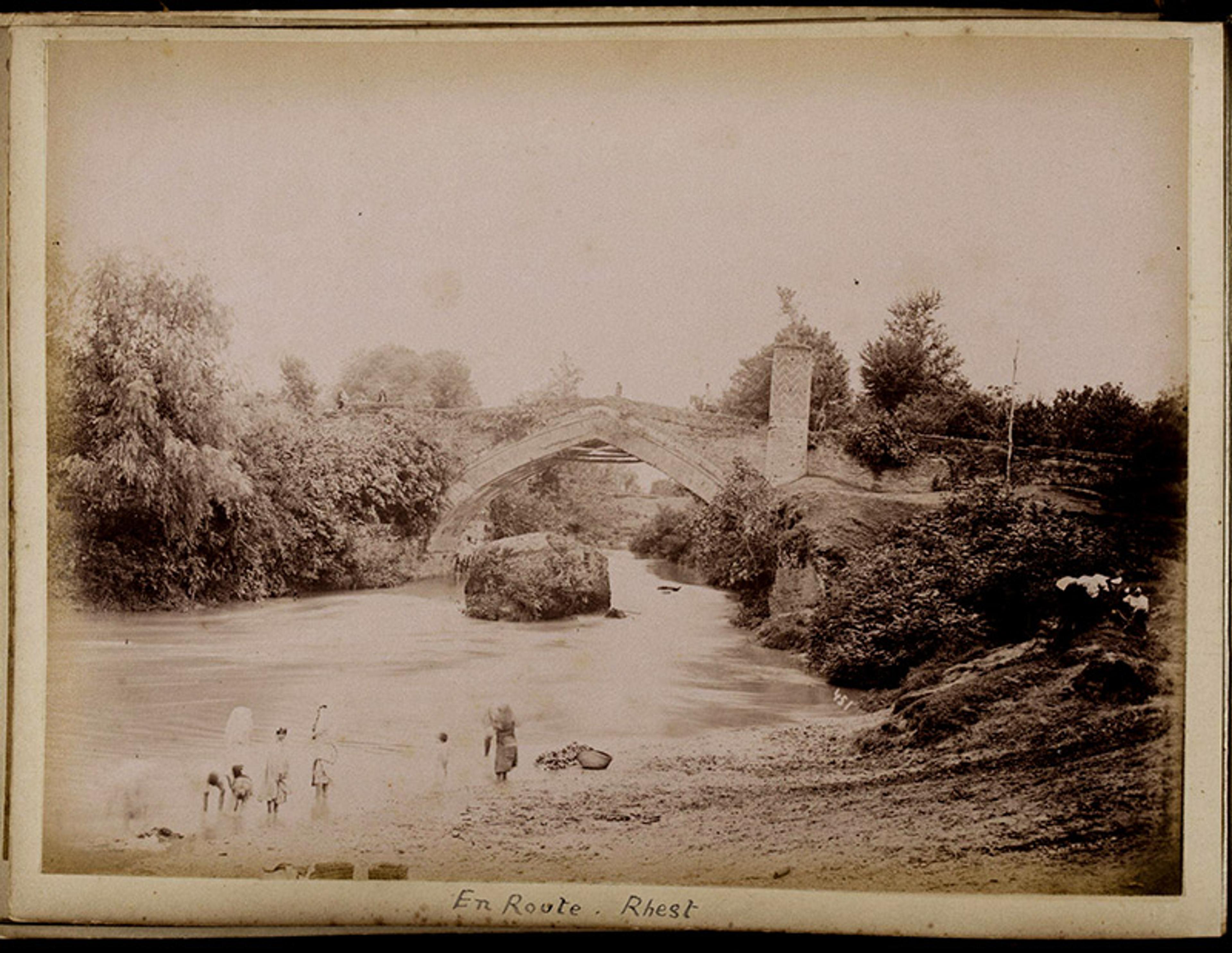 Black-and-white photo of a stone bridge over a river with people on the riverbank, surrounded by trees labelled ‘En Route Rhest’.