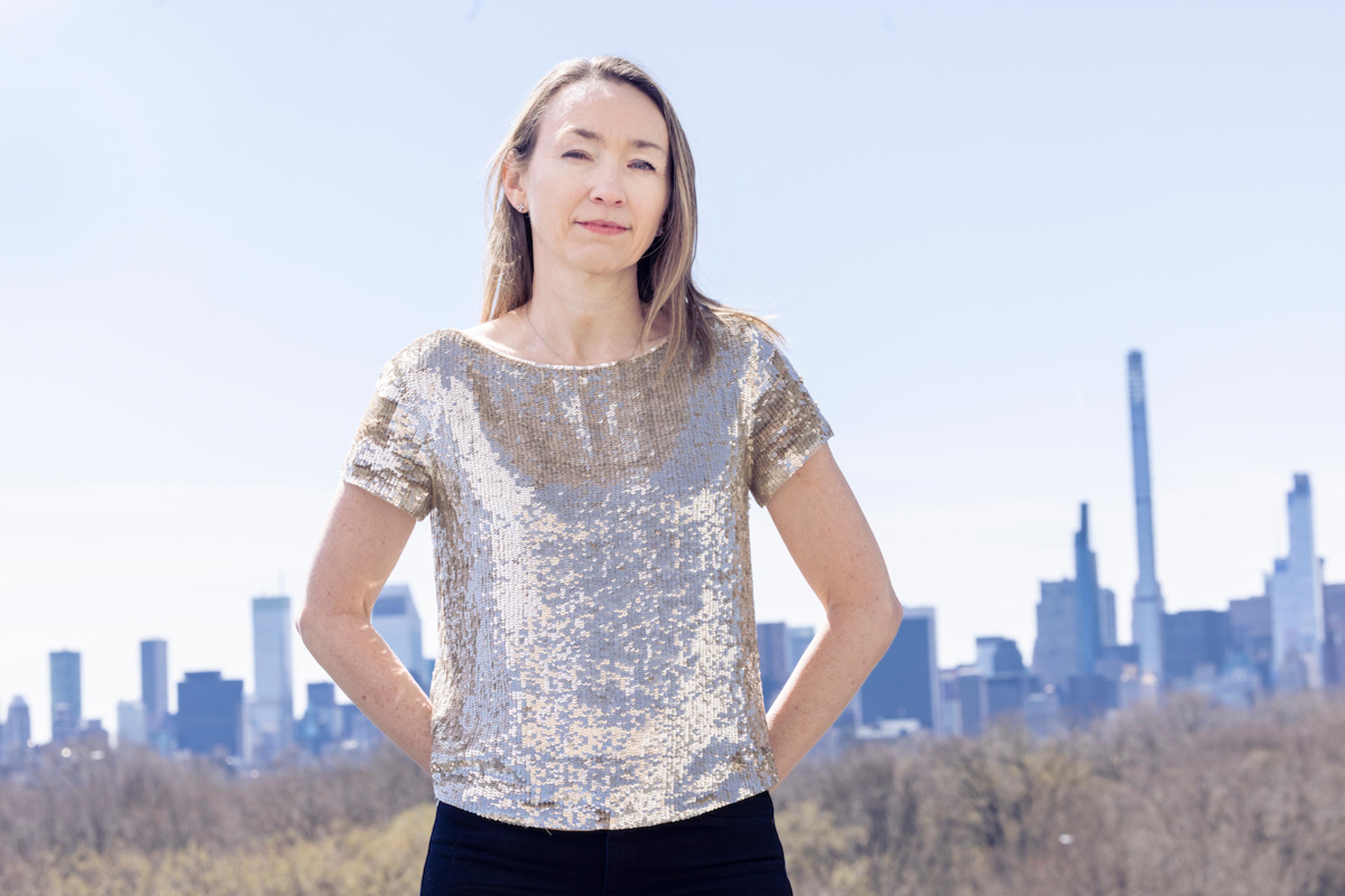 A woman in a shiny silver top standing outdoors with a city skyline in the background on a clear day.