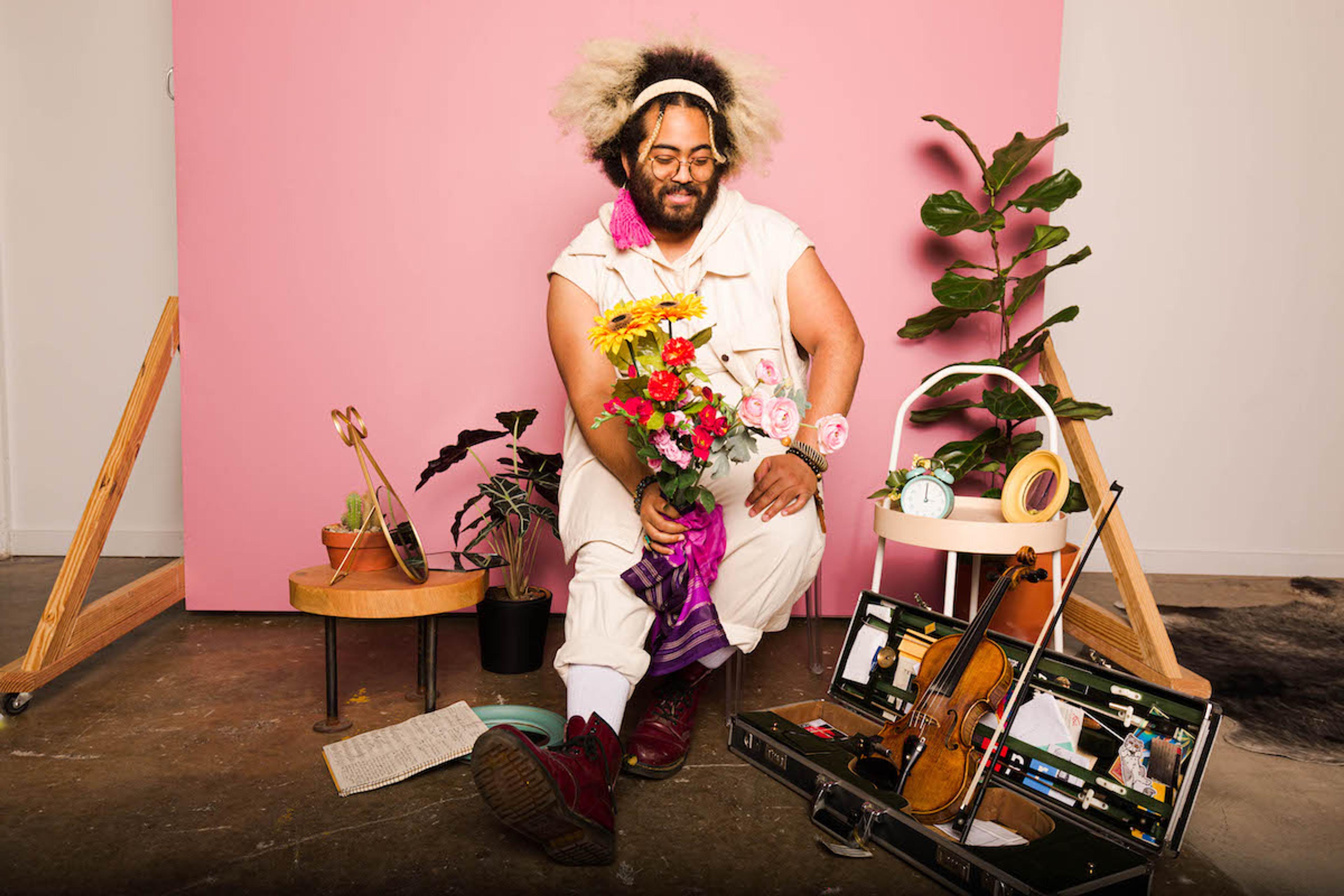 A bearded person in a pale outfit holding flowers, sitting in a studio with plants, a violin case, and a pink background.