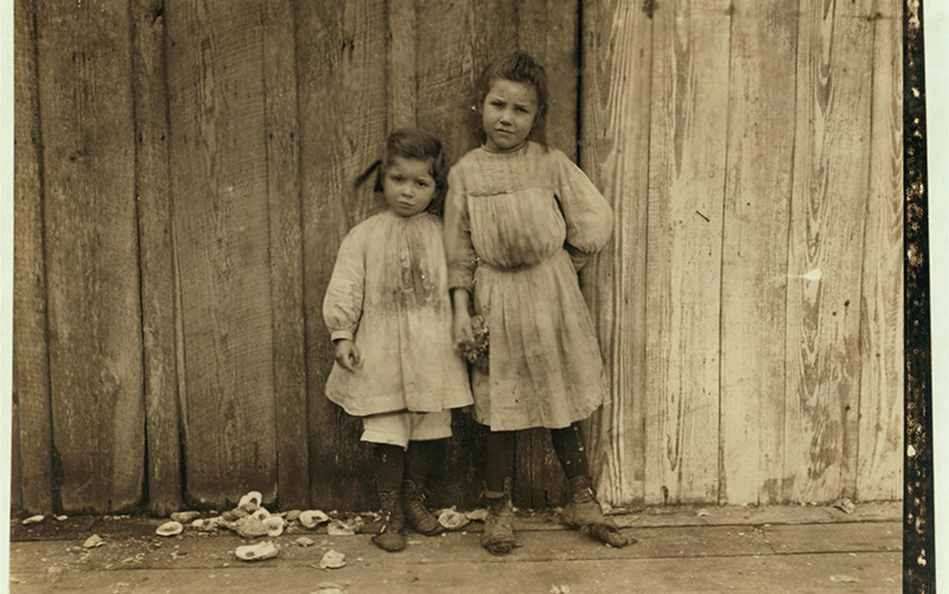 A sepia-toned photo of two young girls standing in front of a wooden wall, both wearing dresses and holding hands, looking at the camera.