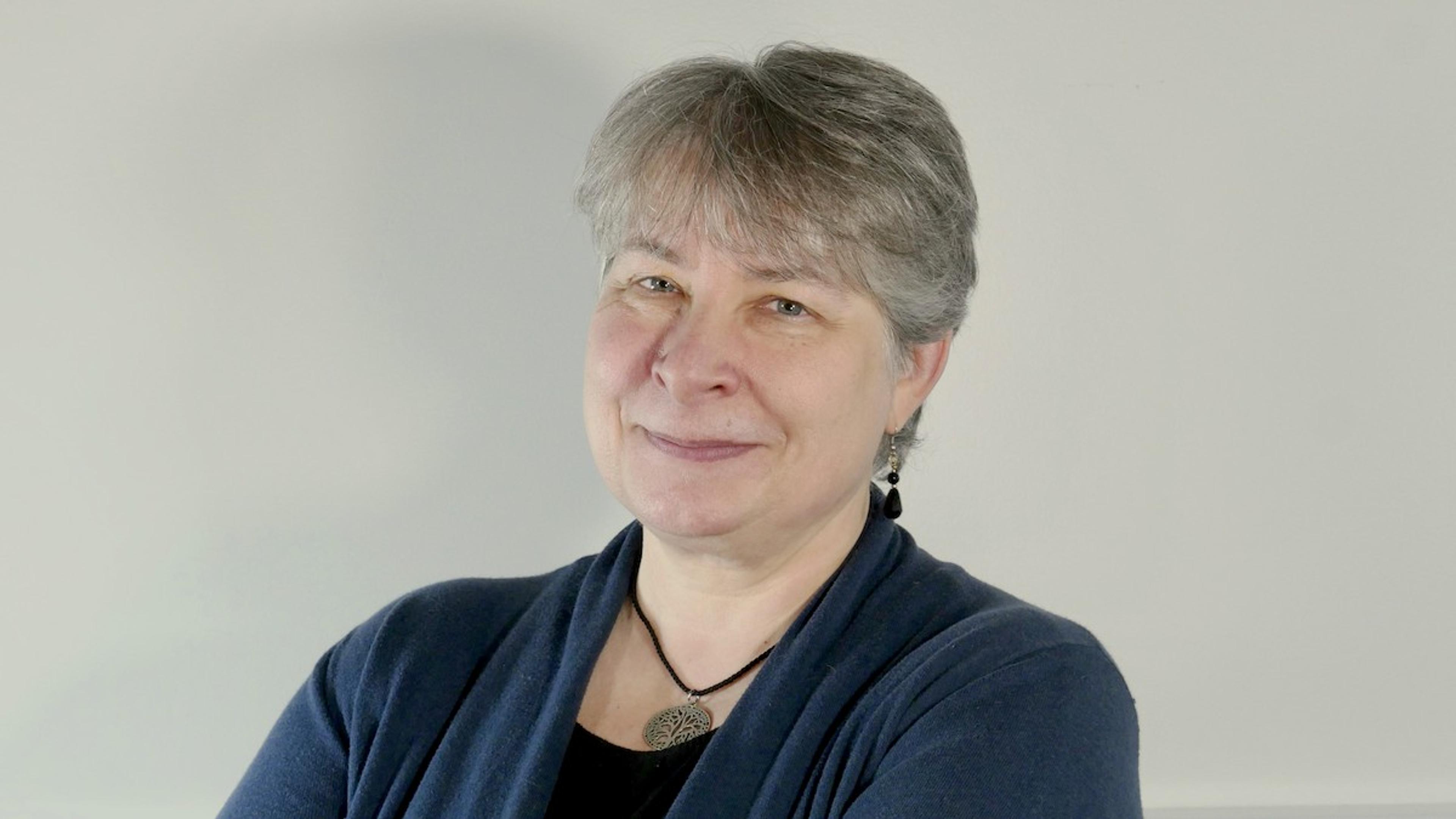 A smiling woman with short grey hair wearing a blue cardigan and necklace standing against a light grey background.