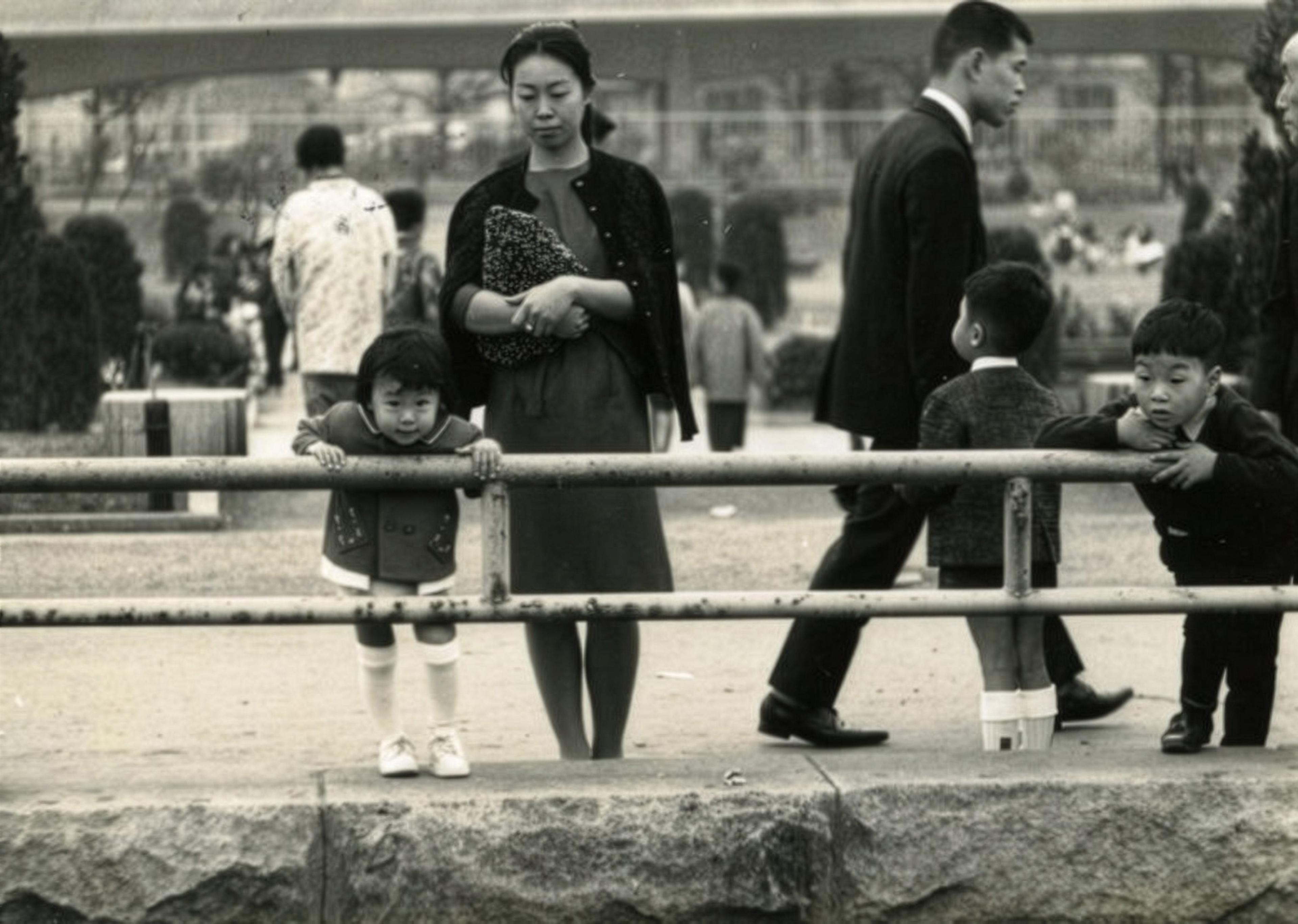 Black and white photo of a woman and children by a railing in a park, with other people in the blurred background.