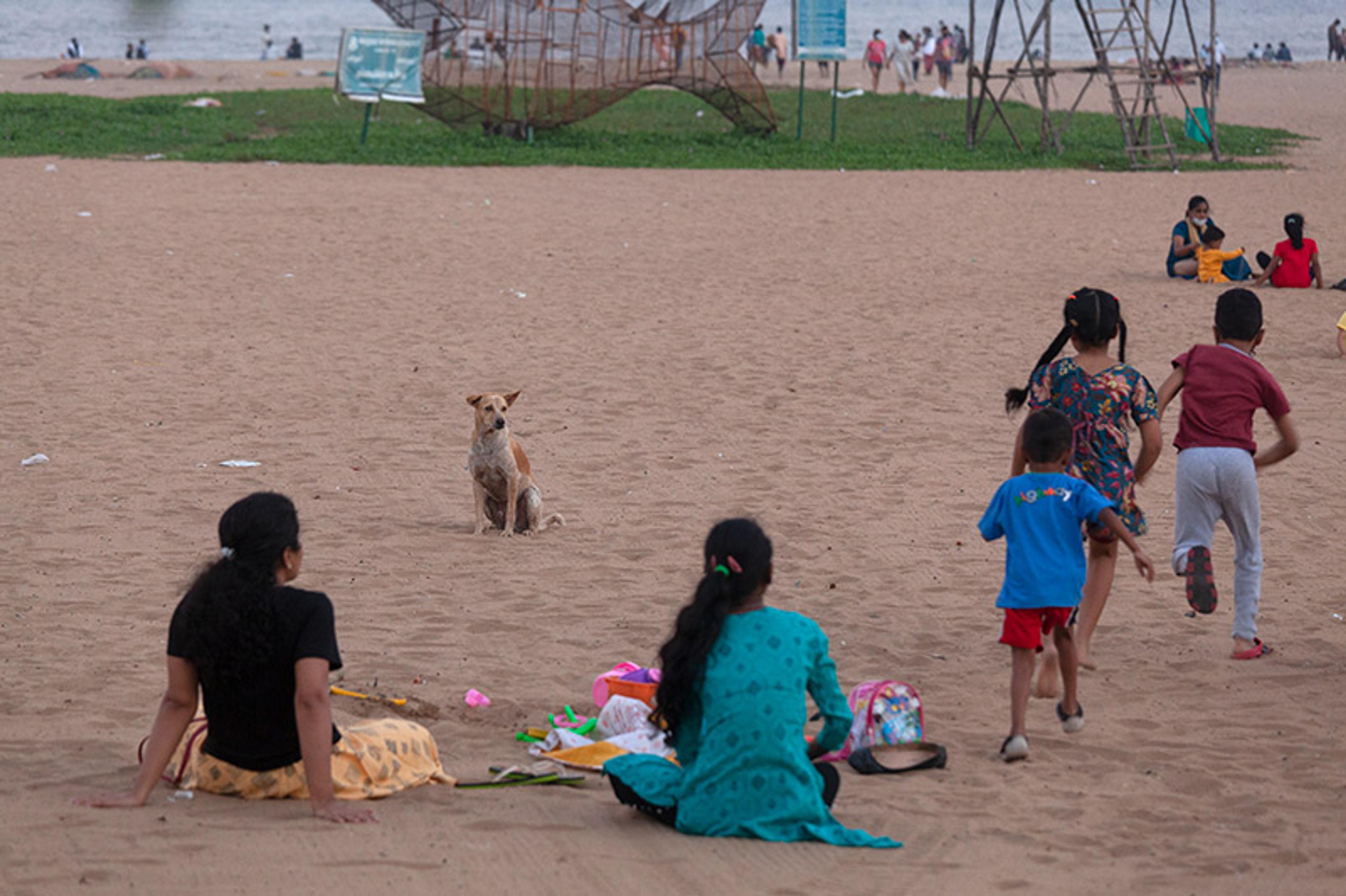 People sitting and children running on a sandy beach, with a dog in the distance and some art structures in the background.