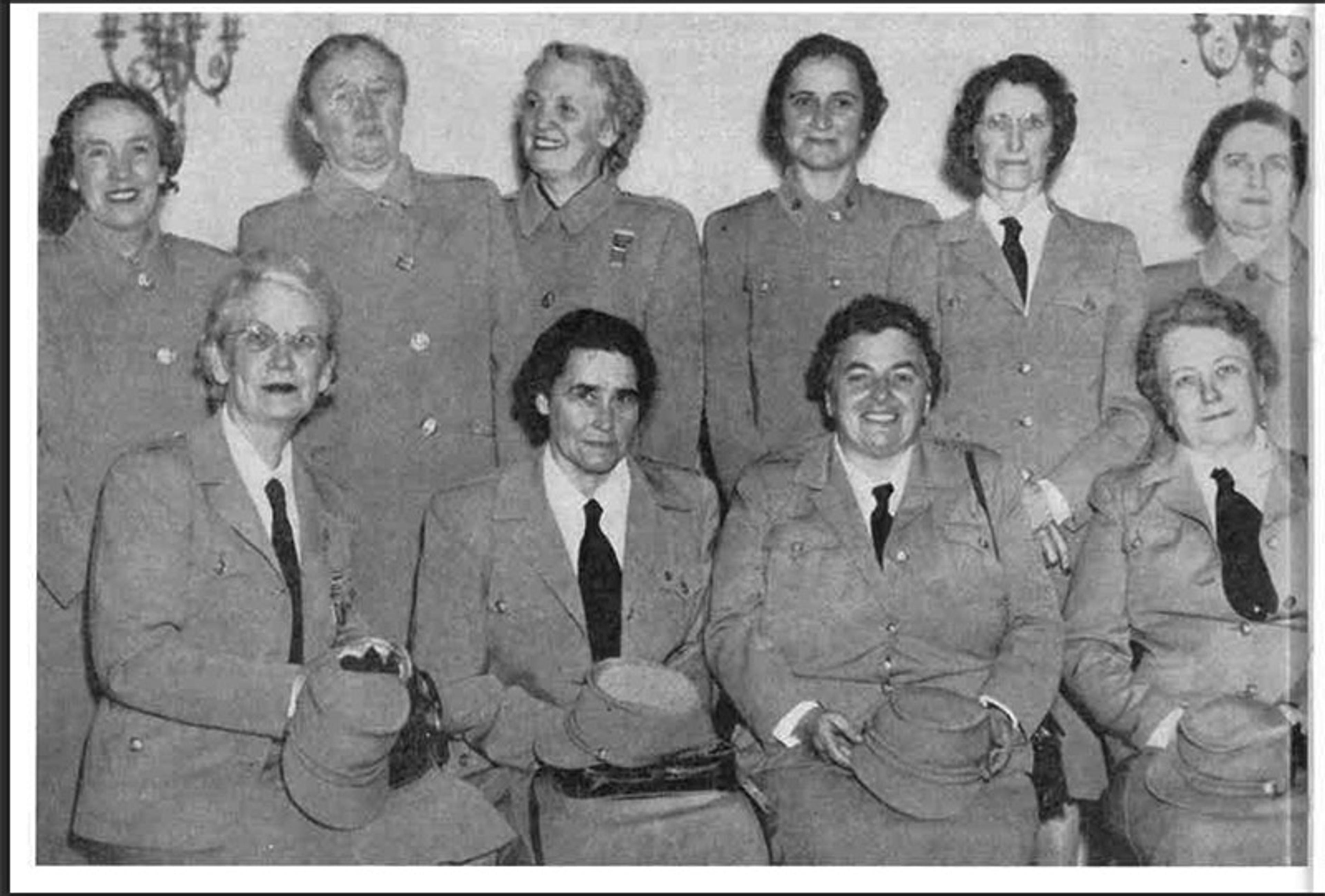 Black-and-white photo of 10 women in uniform, seated and standing in two rows, holding hats, smiling, indoors against a wall.