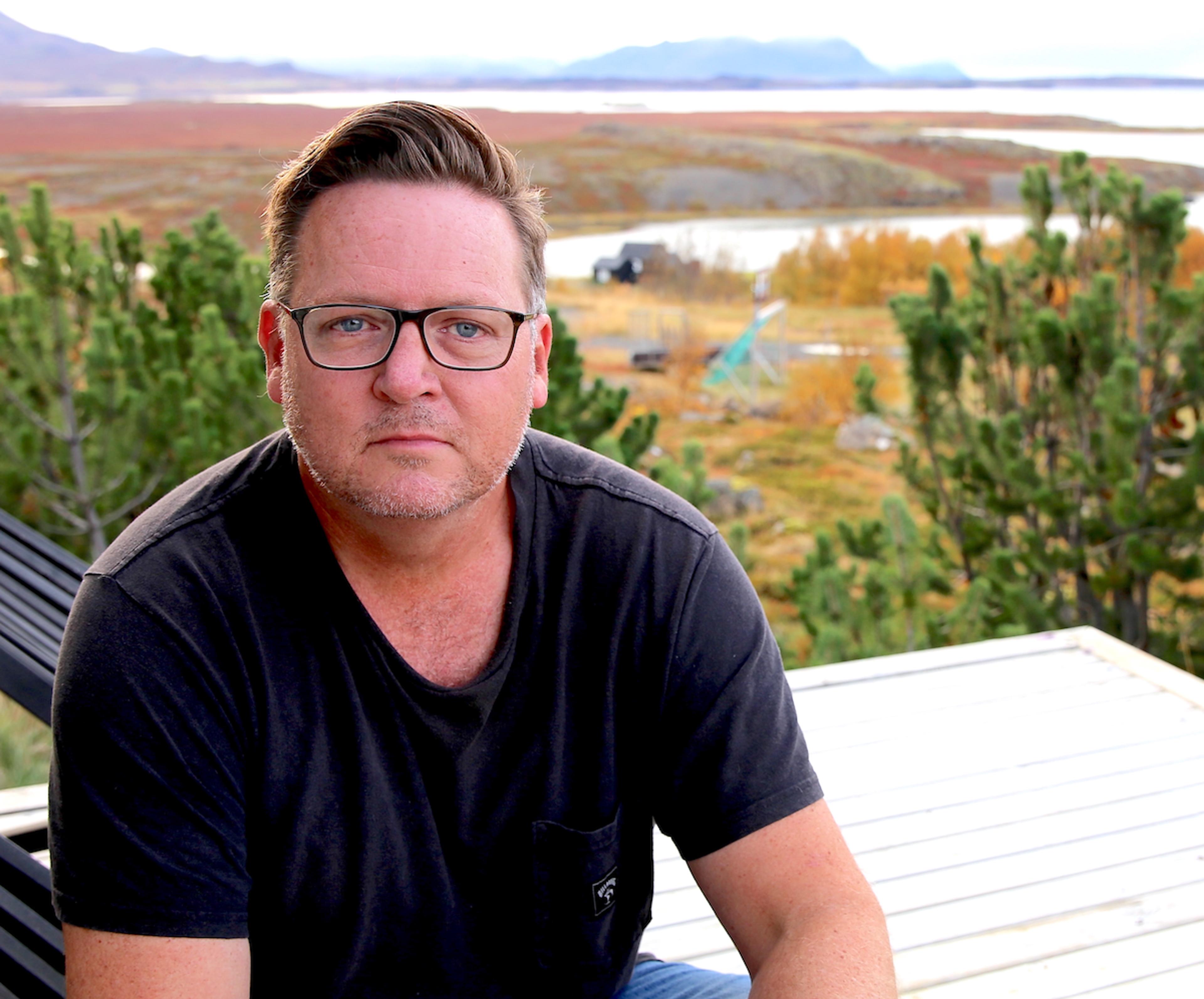 A man in glasses and a black T-shirt on a deck overlooking a scenic landscape with trees, a lake and mountains.