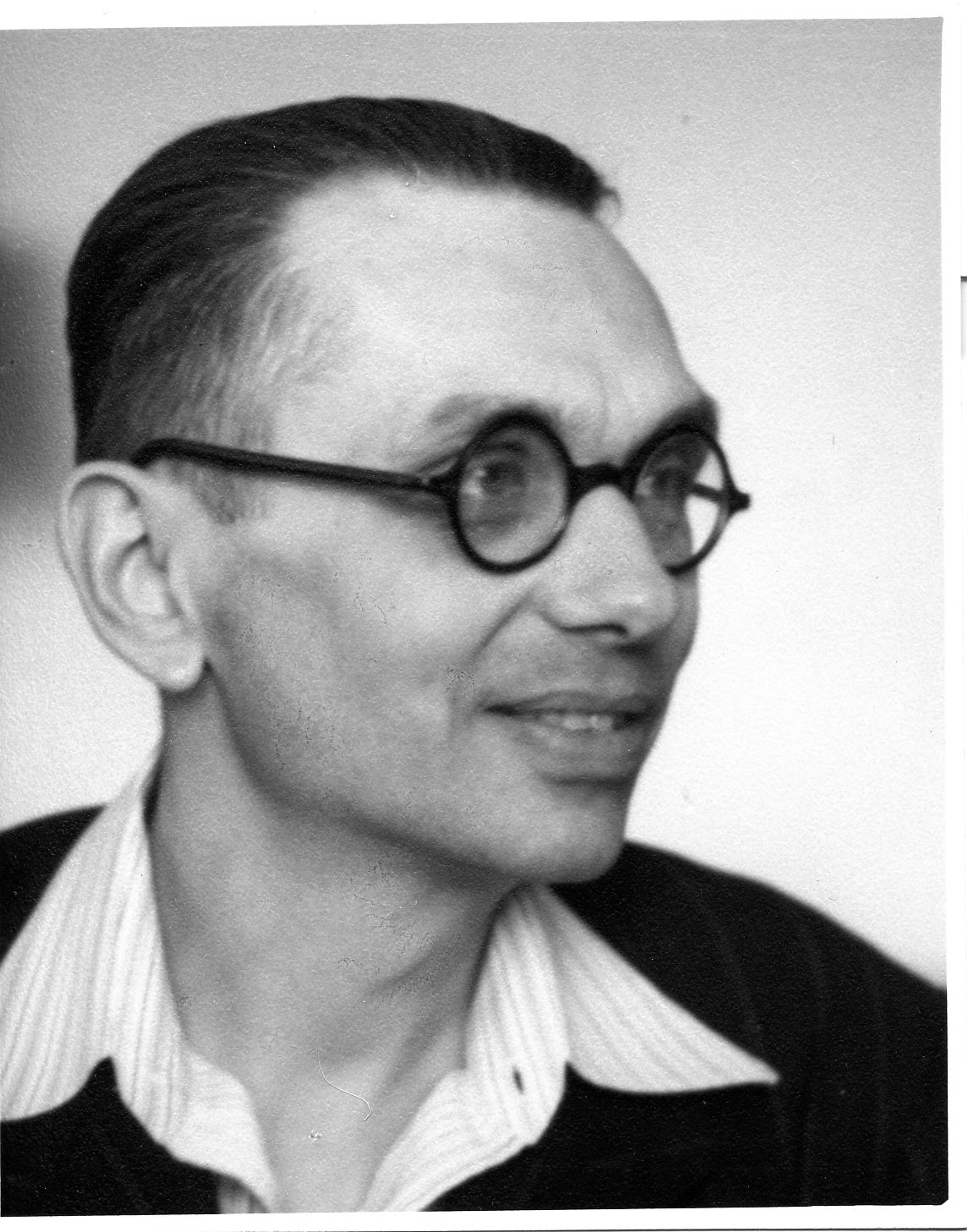 Black and white photo of a man in glasses, wearing a striped shirt and dark jacket, with short hair.