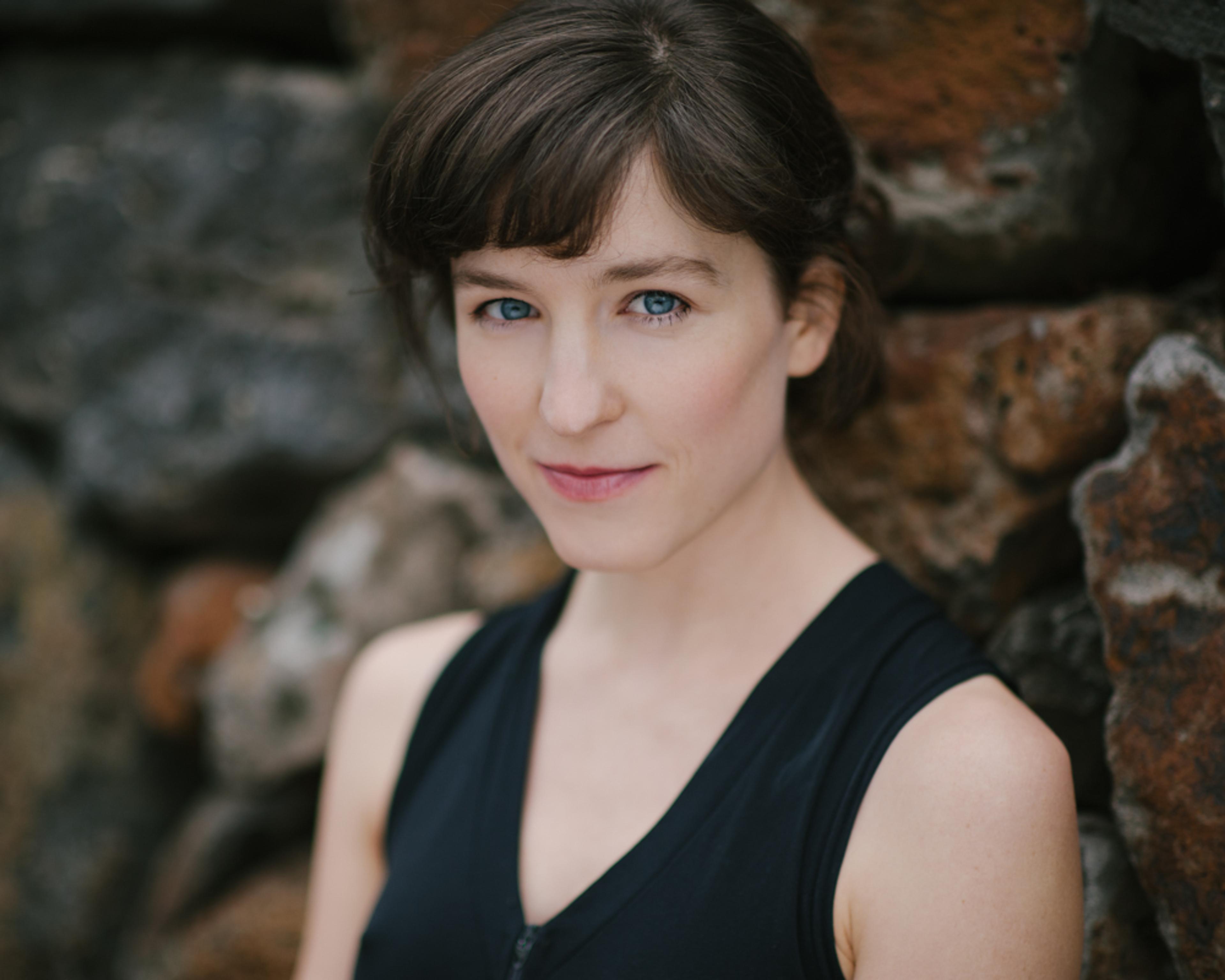 A woman with short brown hair wearing a sleeveless black top, standing in front of a textured stone wall.