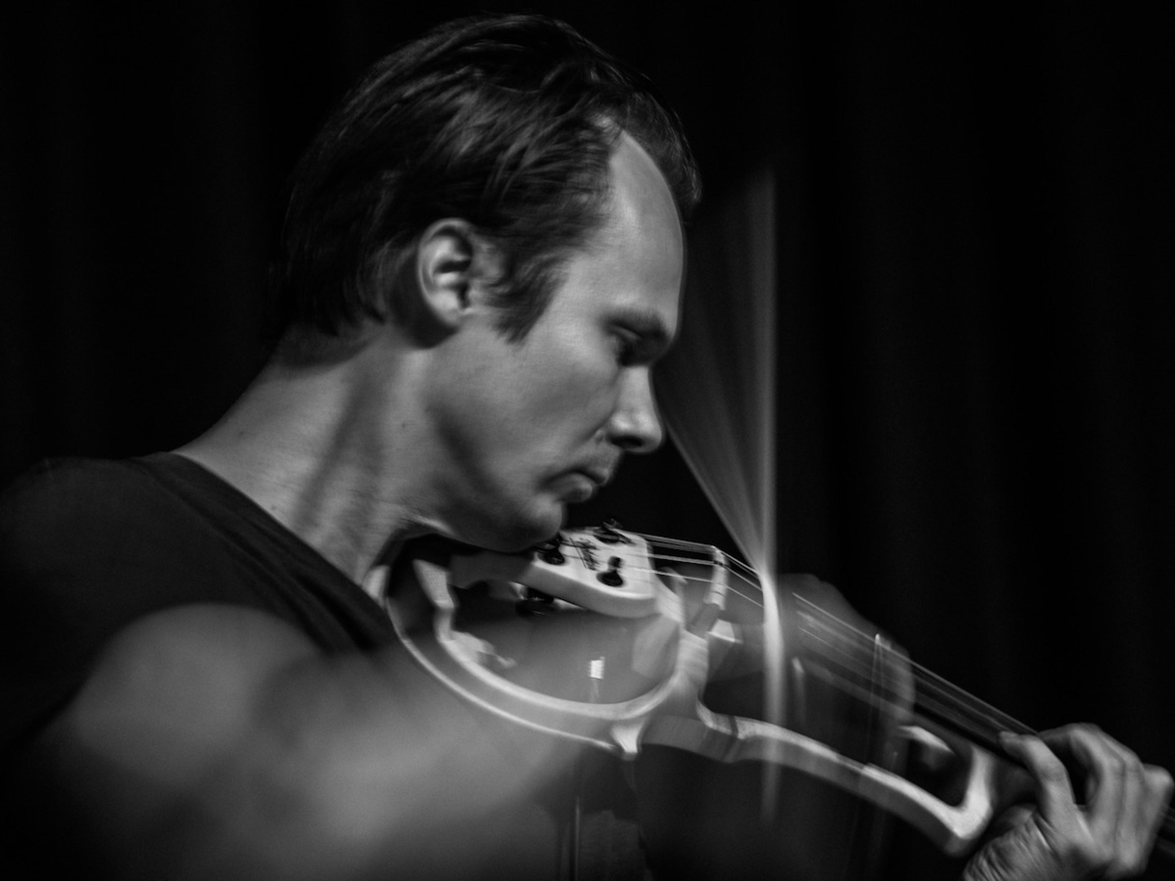 Black-and-awhite photo of a man playing the violin in focus under low light background, his face shows concentrated emotion.