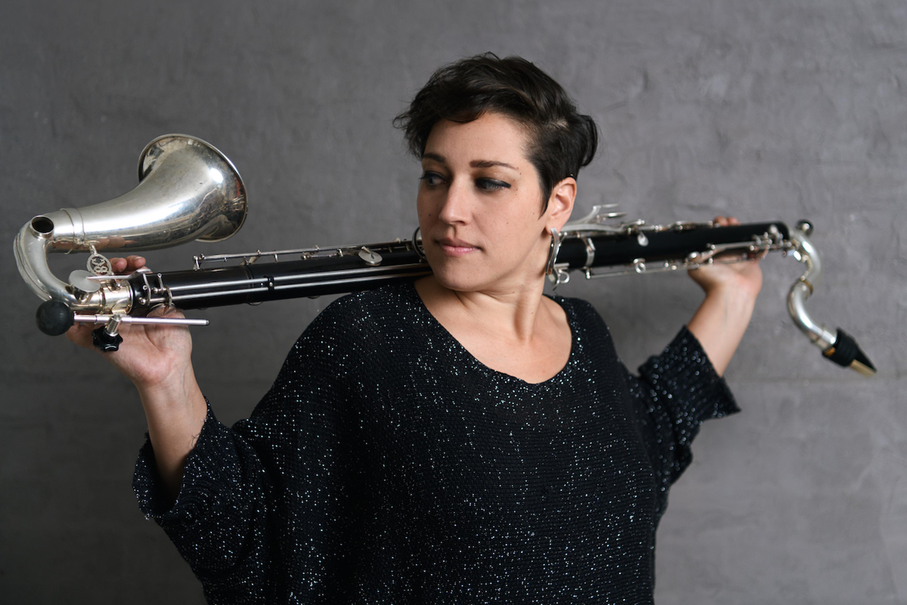 A woman with short dark hair holding a large woodwind instrument over her shoulders against a grey background.