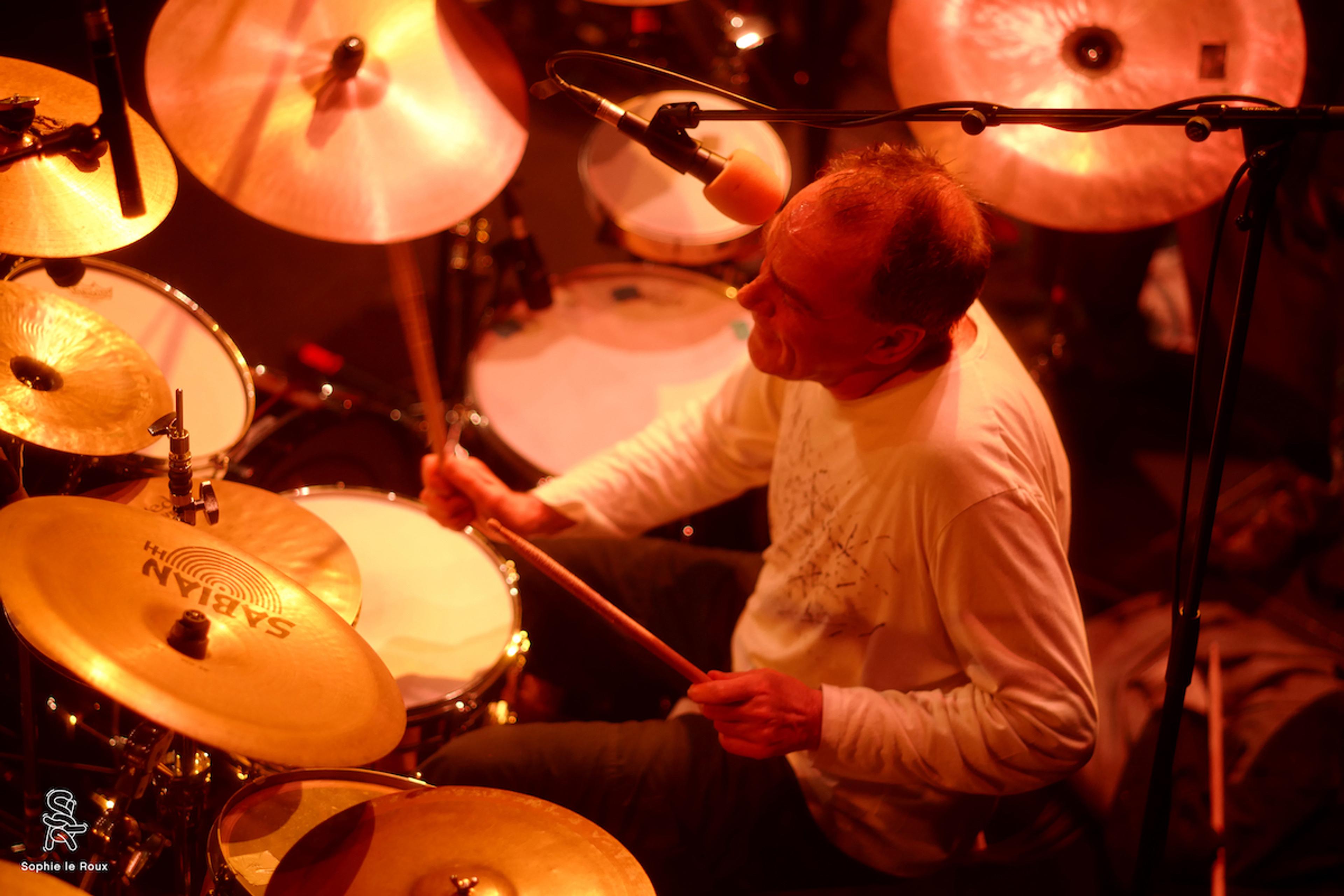 A drummer in a long-sleeve shirt playing a drum kit, illuminated by warm lighting with a microphone overhead.
