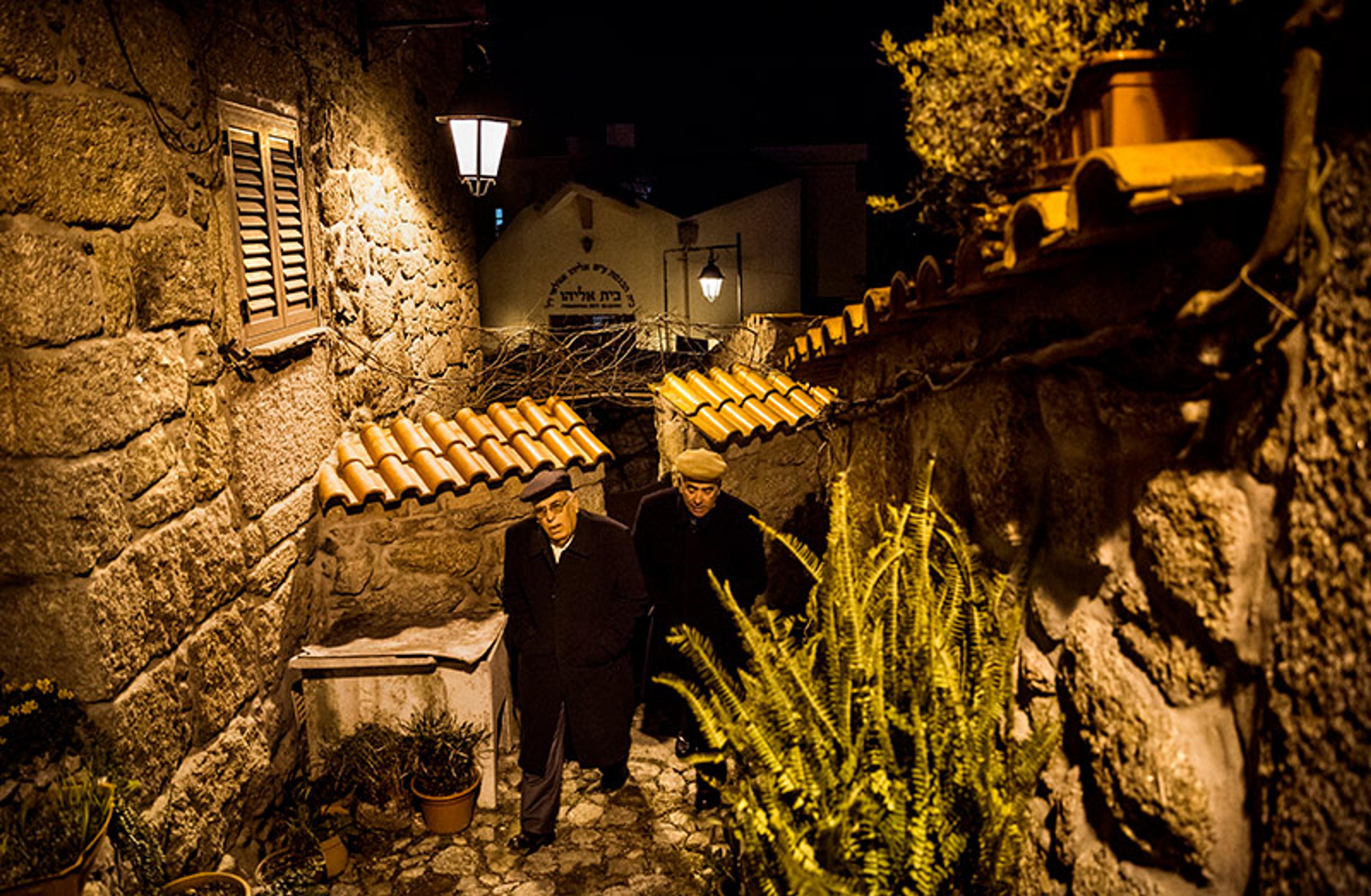 Two elderly men walking down a narrow, dimly lit, stone-paved alley at night, flanked by stone buildings and plants.